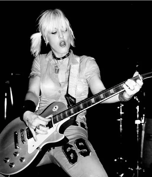 A woman with blonde hair in pigtails playing an electric guitar on stage, dressed in a short-sleeved shirt and shorts, in a black and white photo.