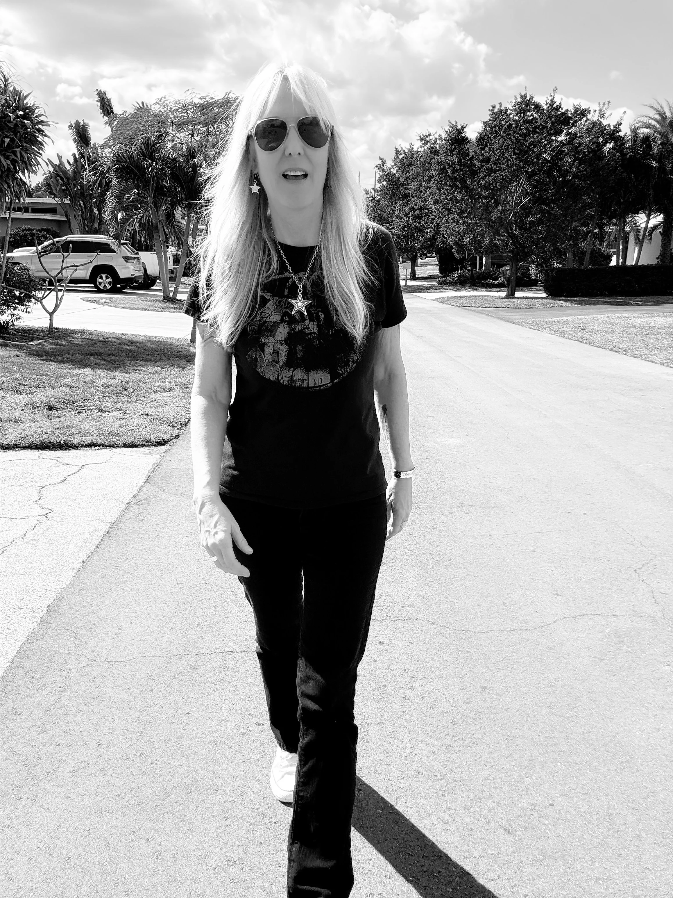 A woman with long, light-colored hair wearing sunglasses, star-shaped earrings, a necklace, a graphic t-shirt, black pants, and white sneakers, walking on a sidewalk in a sunny outdoor setting with trees and parked cars in the background.
