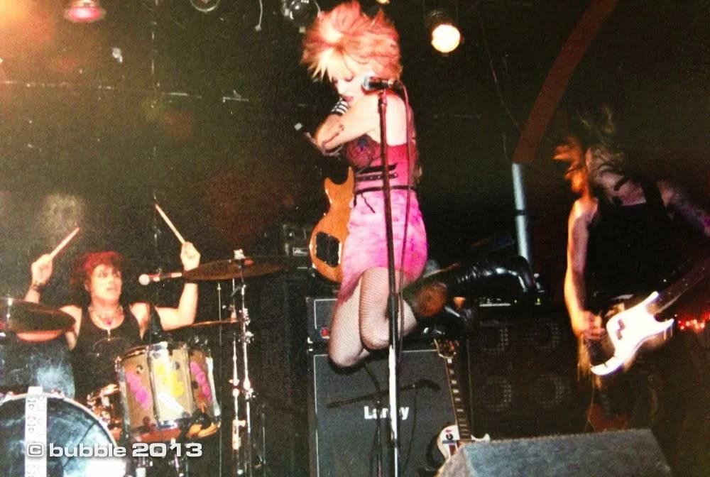 Female vocalist jumping in front of a microphone with a band performing on stage, including a drummer and a guitarist. Stage lights and musical equipment are visible.