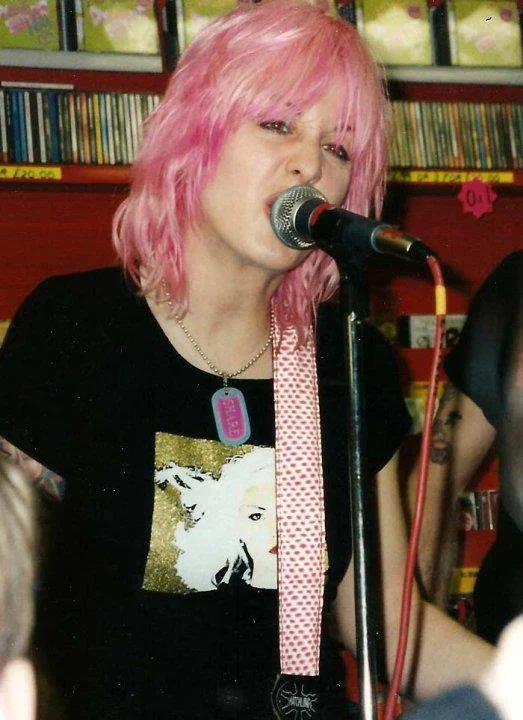 A woman with pink hair singing into a microphone in a record store.