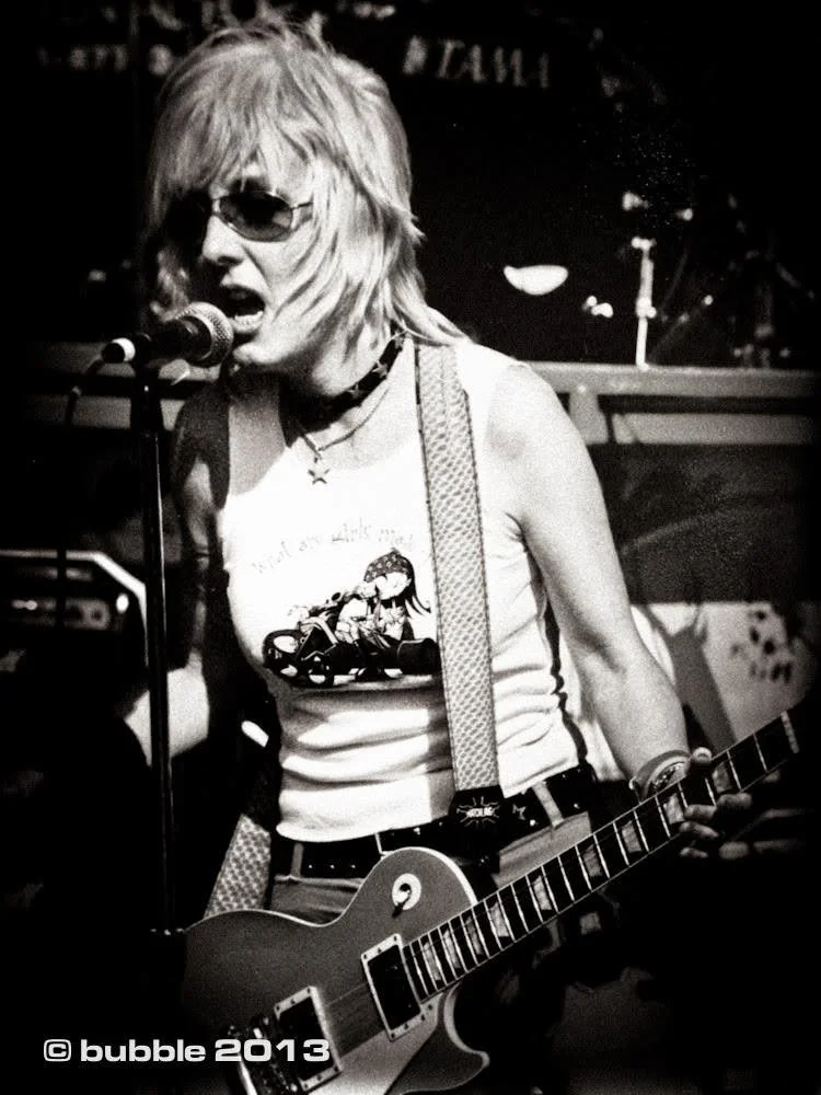 A woman playing an electric guitar and singing into a microphone, wearing sunglasses, a graphic tank top, and a choker, on stage in a live music performance, black and white photo.