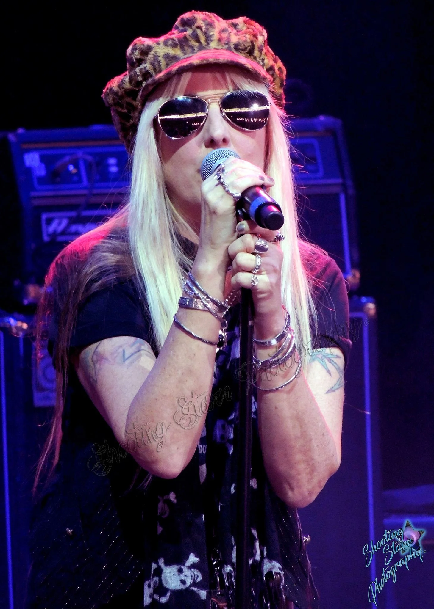 A woman with long blond hair wearing a leopard print beret, round sunglasses, and multiple rings and bracelets, singing into a microphone with an amplifier in the background.