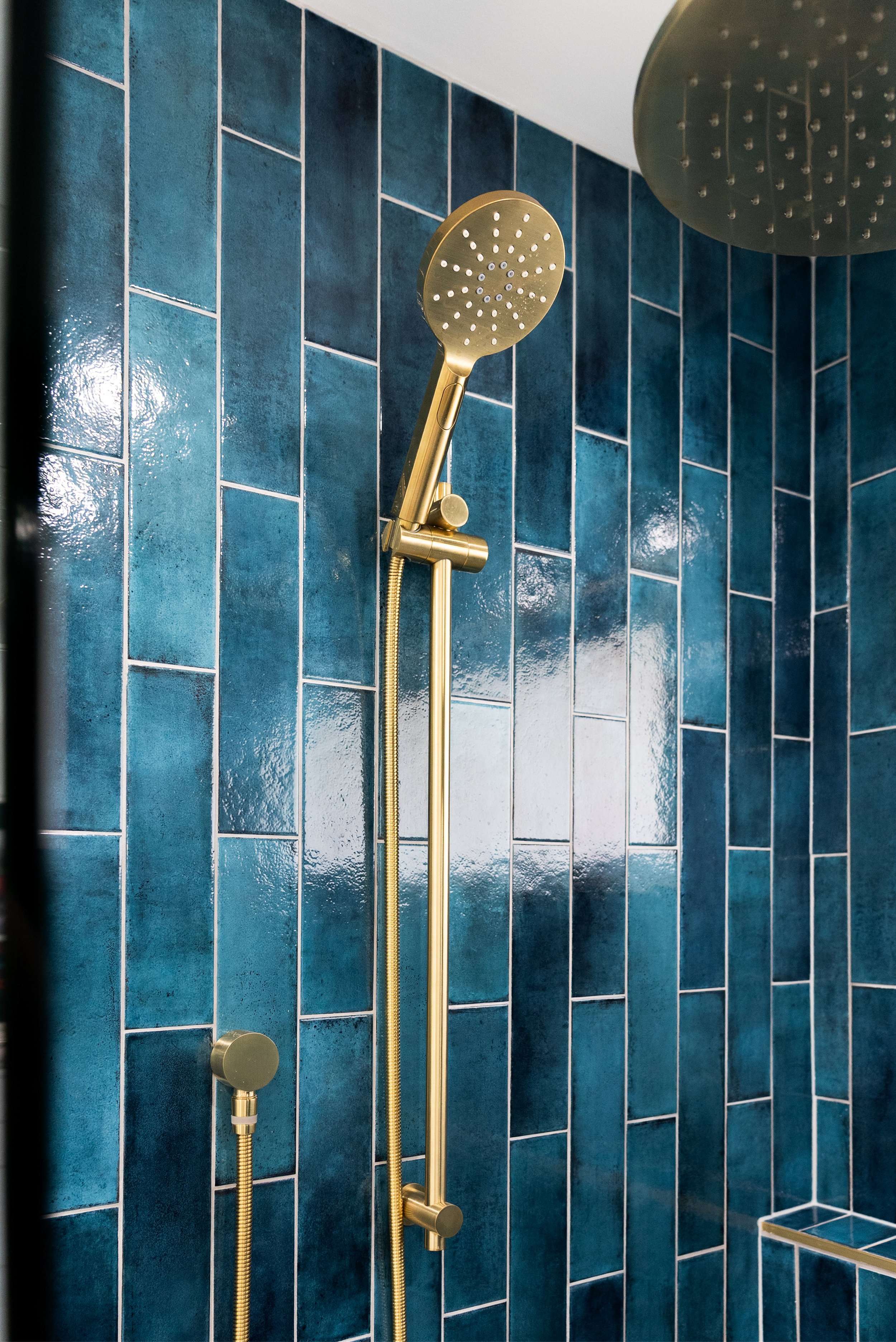 A bathroom shower with blue tiled walls, a large round brass showerhead, and a brass handheld showerhead on a matching brass rail.