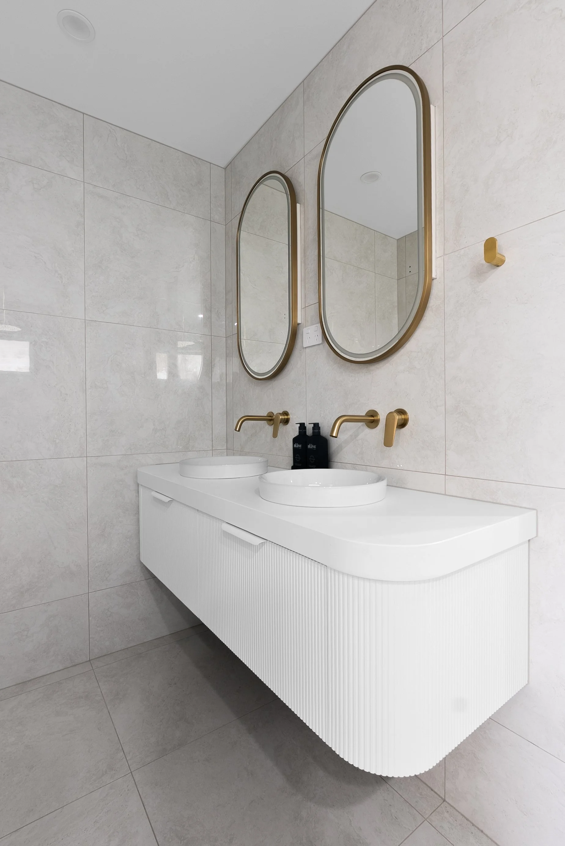 Modern bathroom with a white floating vanity, two oval mirrors with gold frames, two gold wall-mounted faucets, and a light-colored tiled wall.