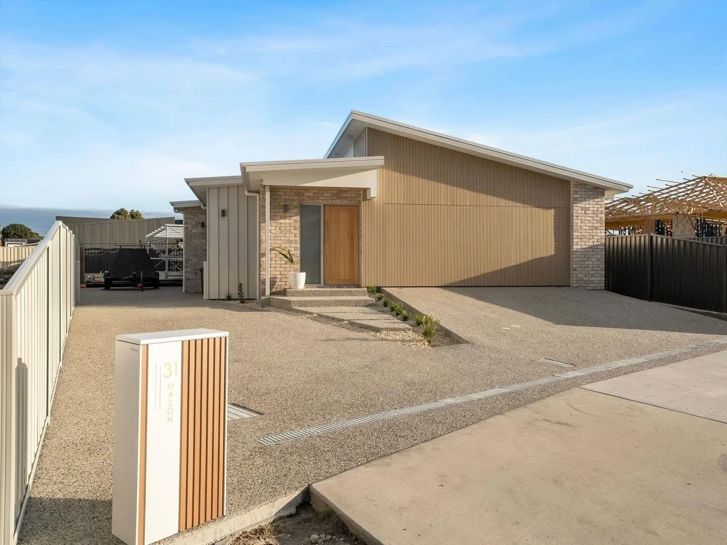Modern single-story house with a minimalistic design, featuring a brick and beige siding exterior, a sloped roof, and a small front porch with a wooden door. The driveway is gravel, with small plants along the pathway, and a modern mailbox in the front yard. There is a partially constructed house or structure visible on the right side of the picture.