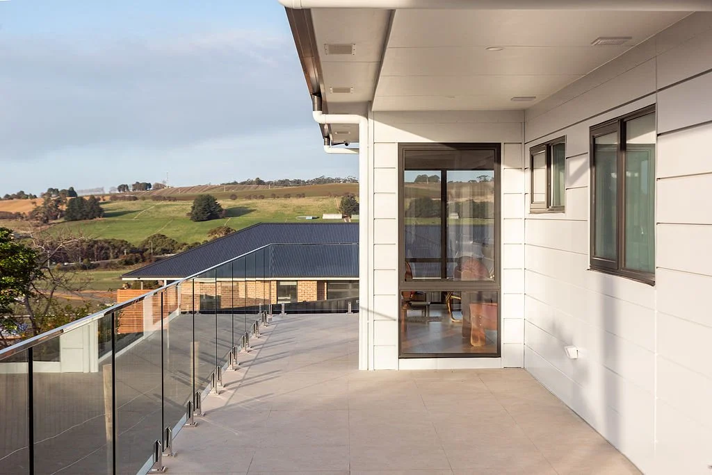 Balcony with glass railing attached to a modern house, overlooking rolling green hills and a neighboring building with a metal roof.