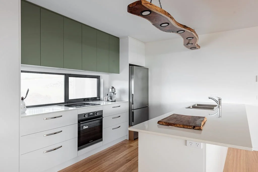 Modern kitchen with white cabinets, green upper cabinets, a black stove, and a stainless steel refrigerator. There is a white countertop island with a cutting board and a sink, a window above the stove, and a wooden ceiling fixture with built-in lights.