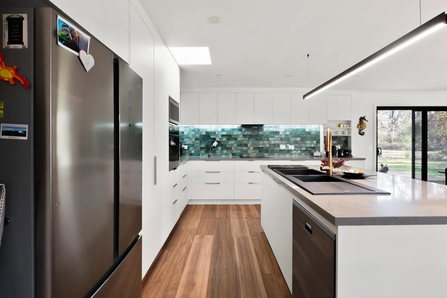 Modern kitchen with white cabinets, a green and blue tiled backsplash, stainless steel appliances, a large island with a sink, and large sliding glass doors opening to an outdoor view.