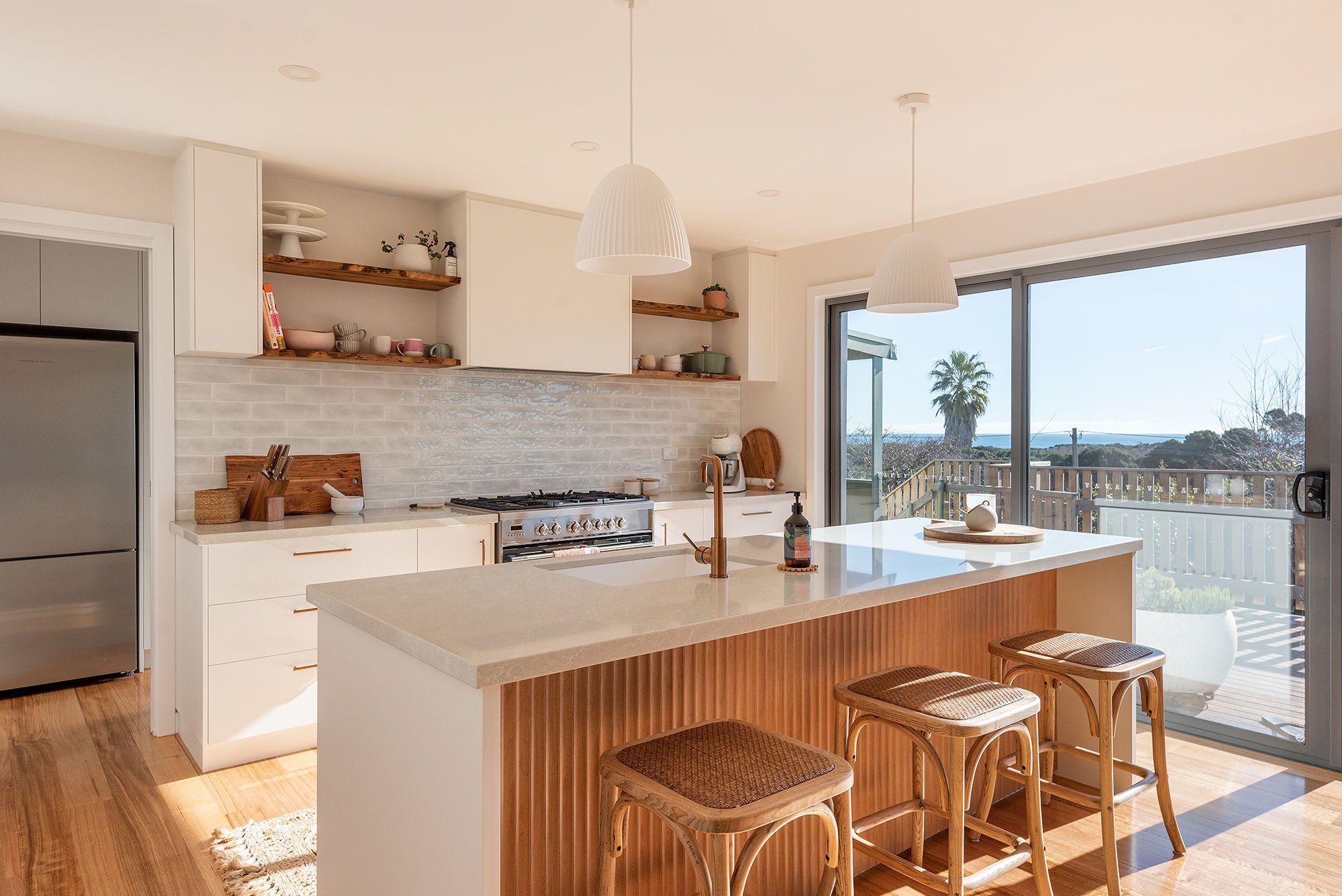 Modern kitchen with white cabinets, wooden open shelves, a white island with wooden paneling, and a view of a deck with a palm tree outside large sliding glass doors.