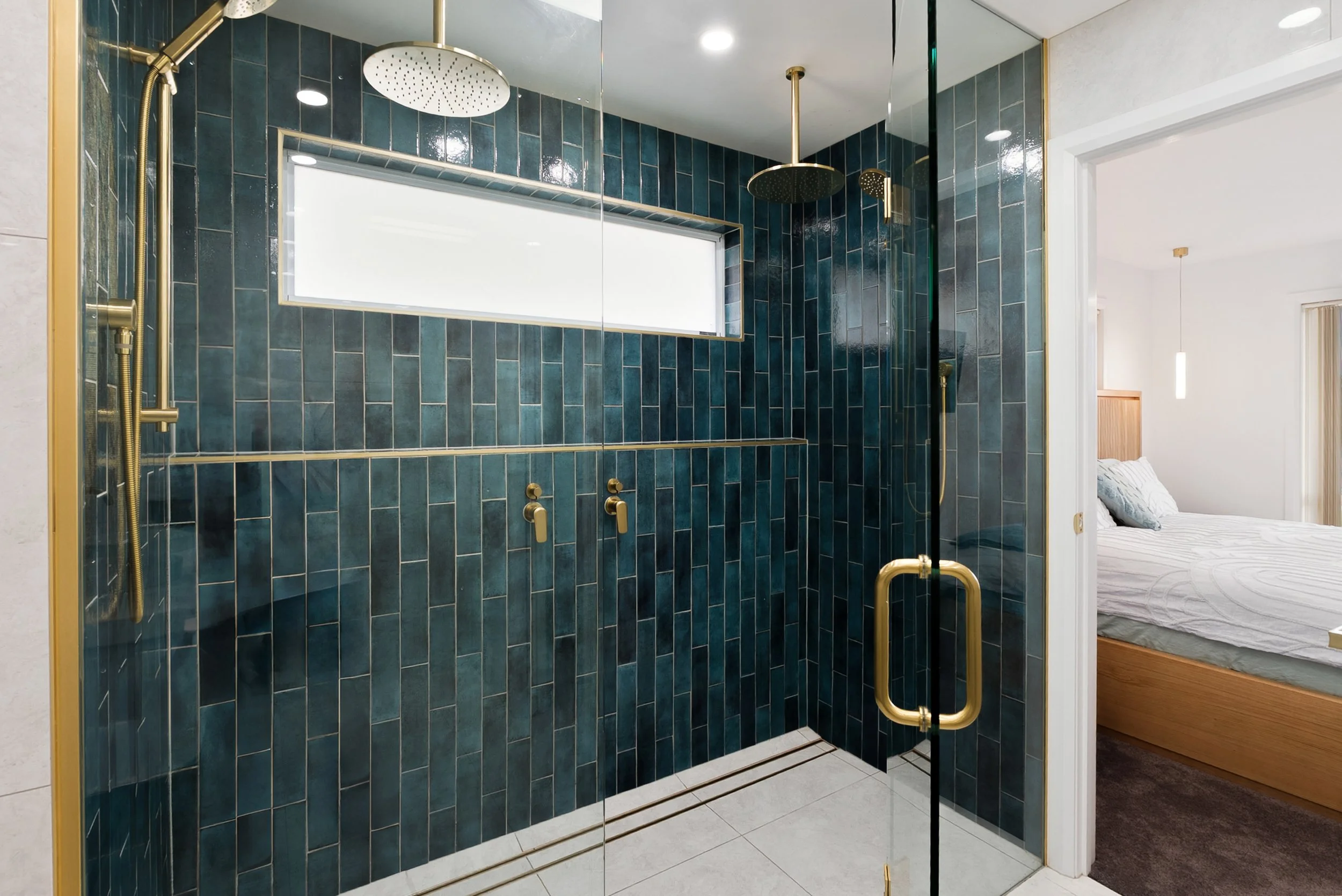 A modern bathroom with a glass shower enclosure featuring dark green rectangular tiles, two rainfall shower heads, and gold fixtures. A narrow horizontal window provides natural light, and part of a bedroom with a bed and pendant light is visible through the doorway.