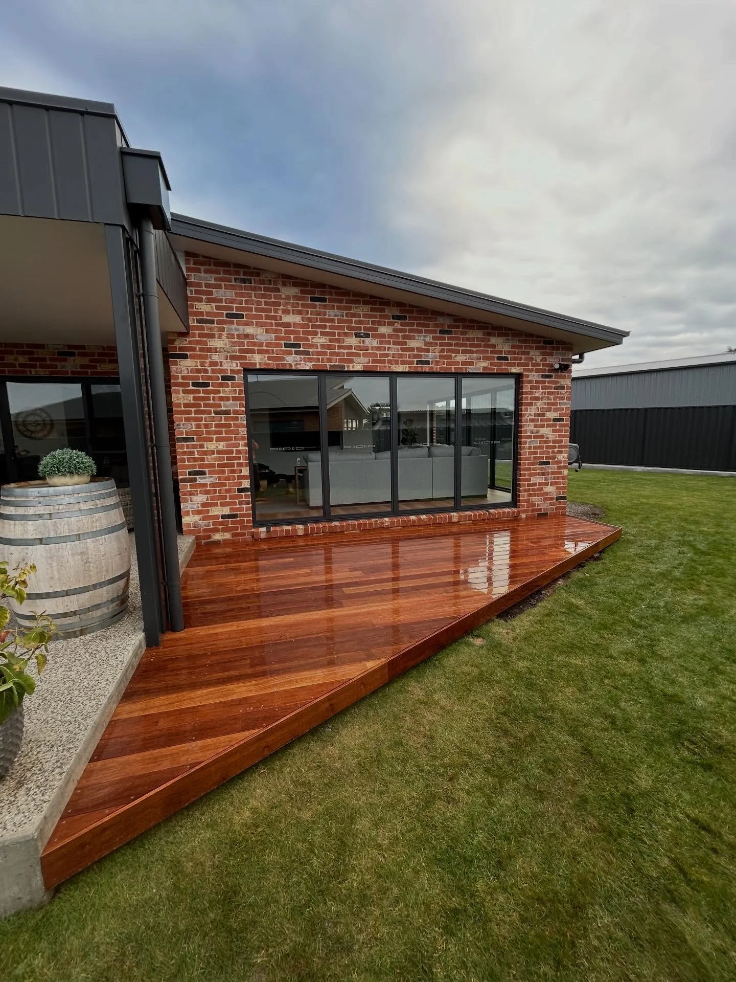 A wooden deck extends from the brick house with large glass sliding doors. The deck is shiny from recent rain. There is a grassy yard surrounding the deck, and a metal fence in the background. The house has modern design elements with brick and metal siding.