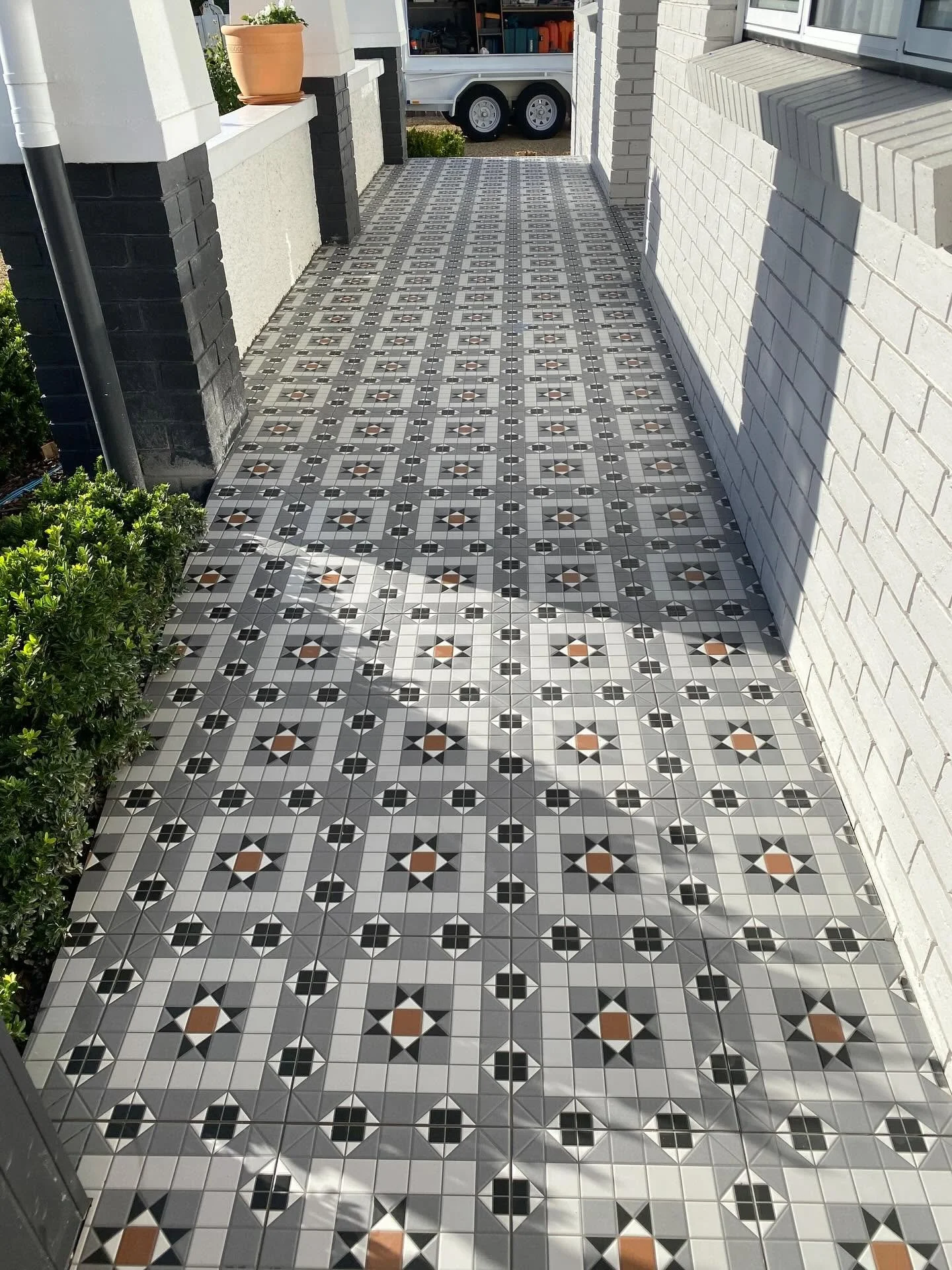 A porch with black and white patterned tile flooring leading to a white brick house. There are green bushes on the left side and a pot with plants on a ledge on the left. In the background, a trailer with tools and a white door are visible.