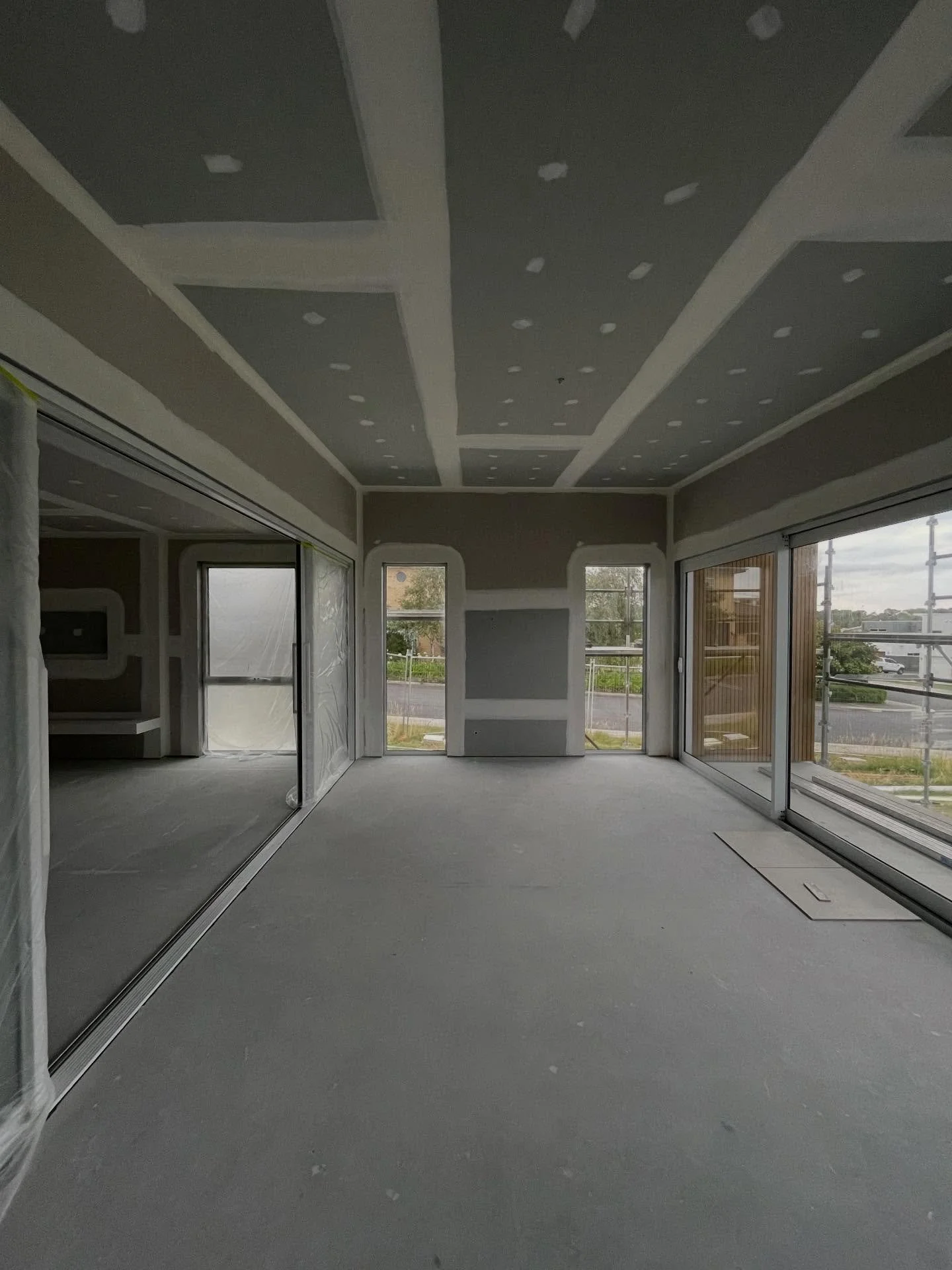 An unfinished room with large floor-to-ceiling windows on one side, showing an outdoor construction site with scaffolding, and a sliding glass door on the other side. The ceiling is partially finished, with drywall and joint compound patches. The floor is bare, gray concrete.