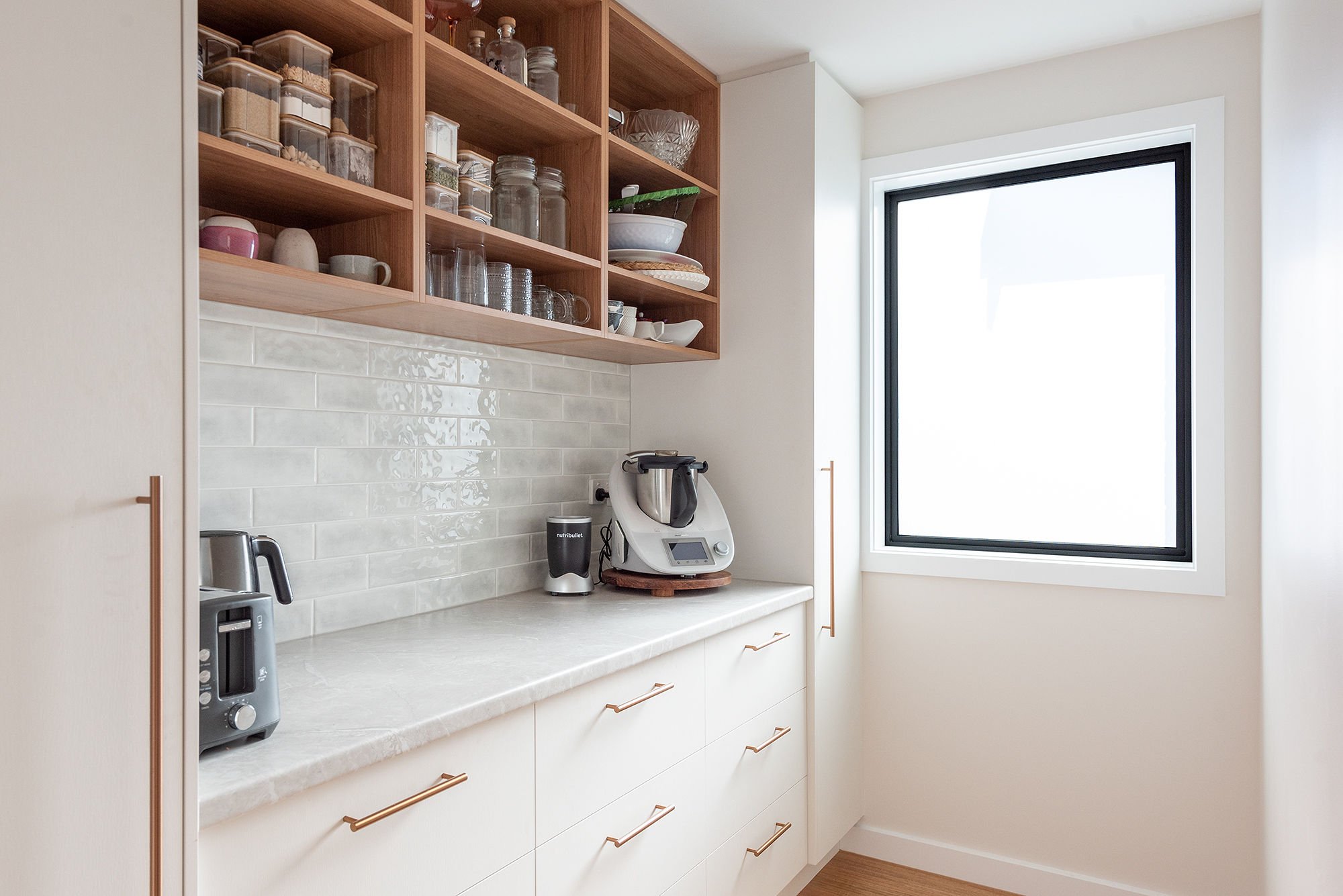 Kitchen countertop with appliances, cabinets, open wooden shelves filled with glass jars and dishes, large window, tile backsplash.