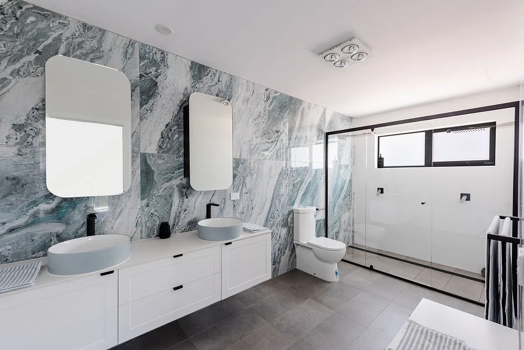 Modern bathroom with marble wall tiles, two white vessel sinks with black faucets, large oval mirrors, a toilet, a glass shower enclosure, and a window with natural light.