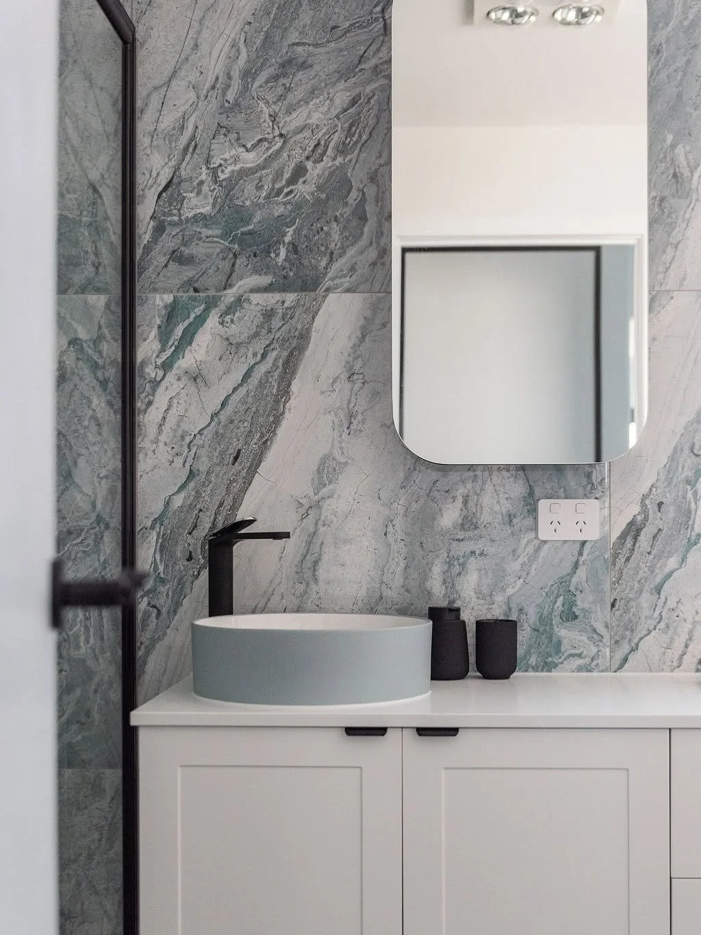 Modern bathroom with marble wall, a white vanity with a circular vessel sink, black faucet, a rectangular wall mirror, two black containers, and an electrical outlet.