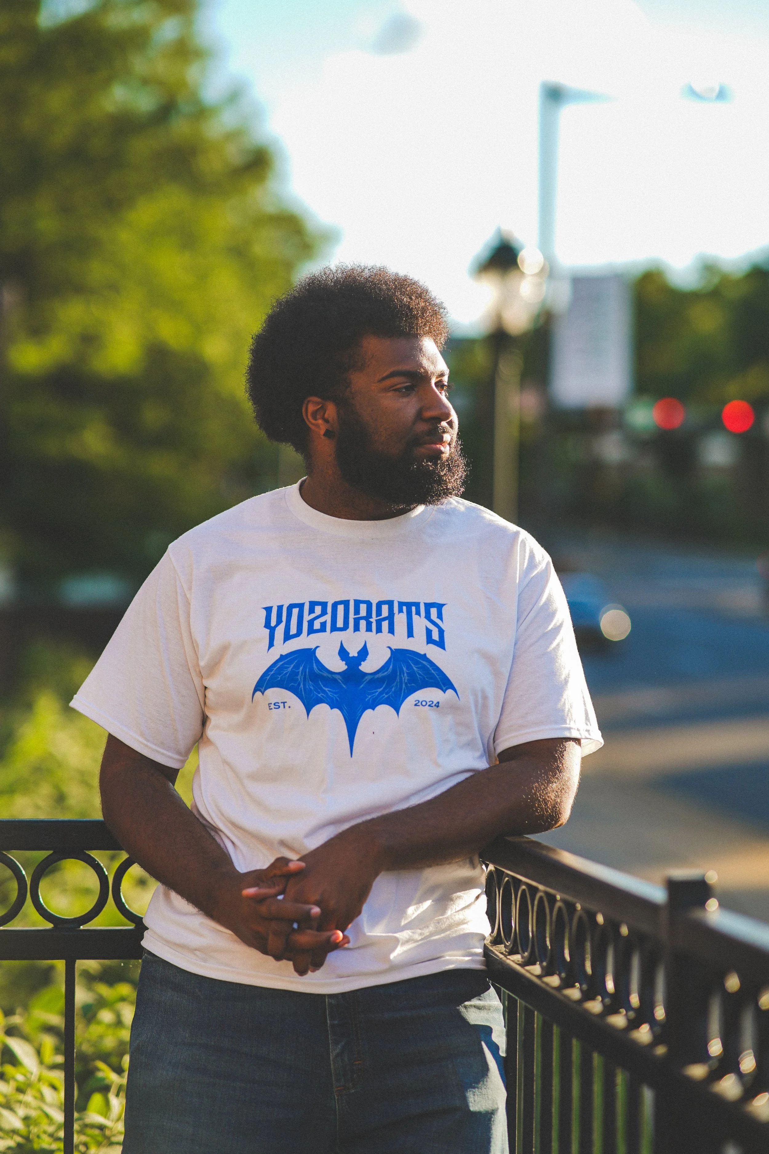 A man with dark curly hair and a beard, wearing a white T-shirt with blue graphic design and text, standing outdoors near a black metal railing, with a background of trees, a blurred sign, and traffic lights under a clear sky.