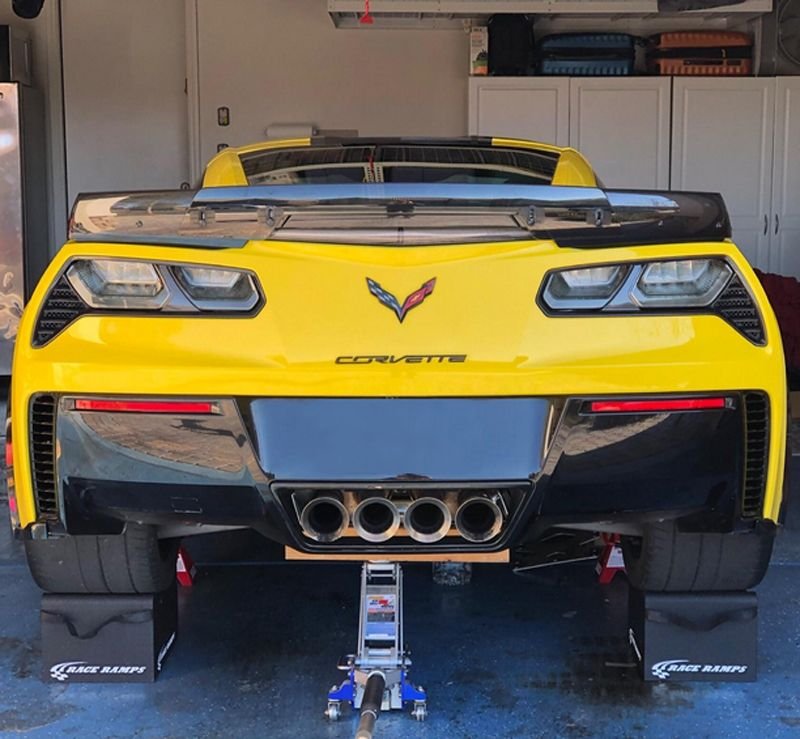 Yellow Chevrolet Corvette car on jack stands in a garage, viewed from the front, with quad exhaust pipes and a Corvette emblem.
