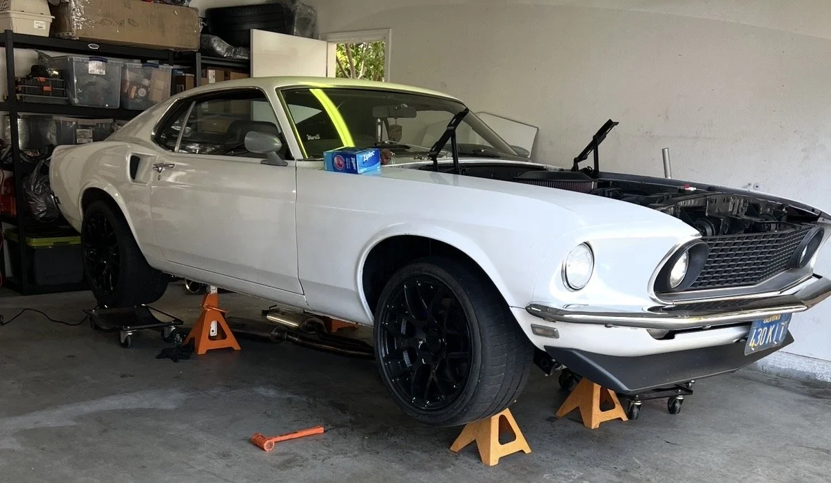 A white vintage car on jack stands in a garage, with its front hood open and a box on the passenger seat.