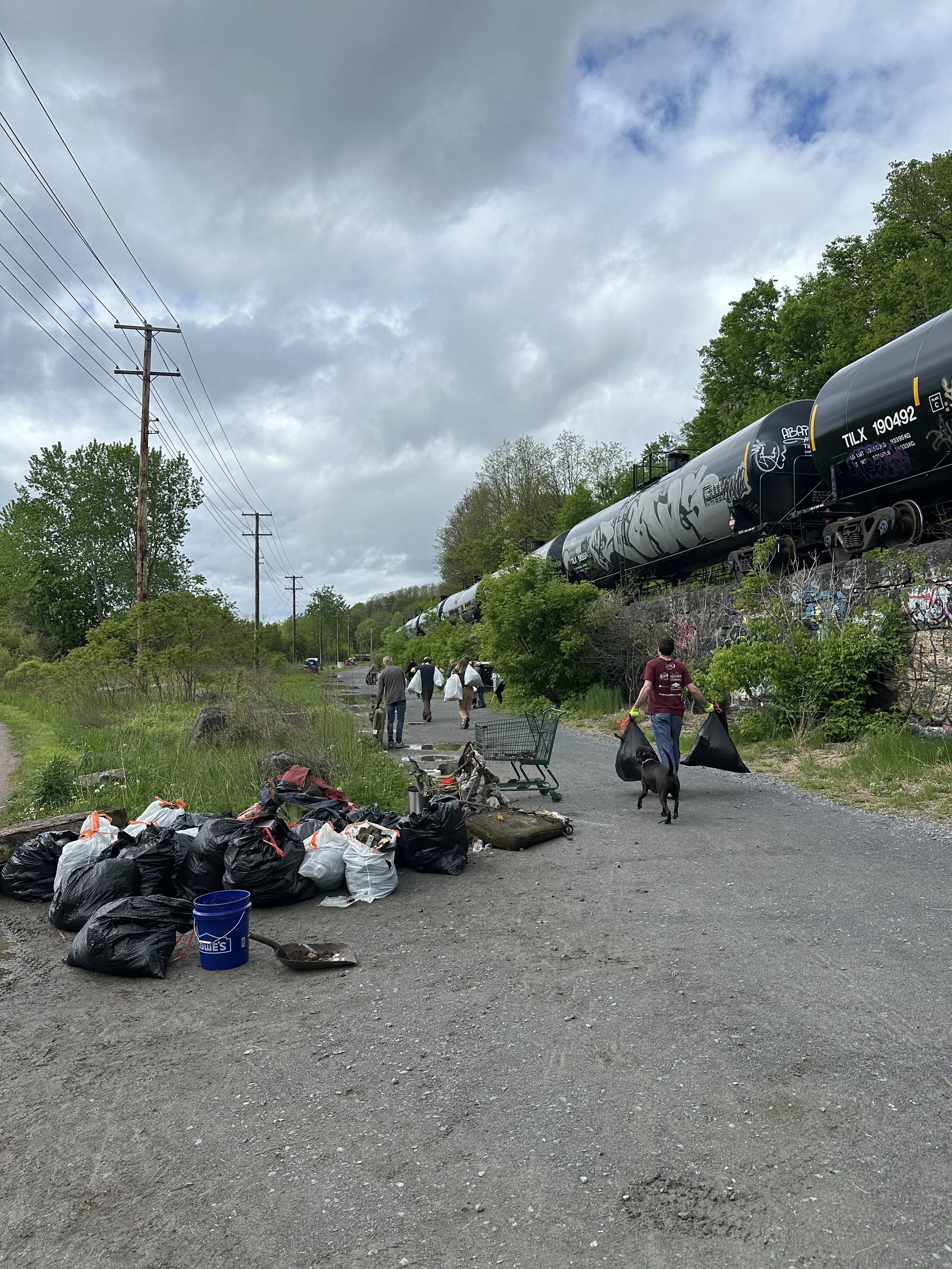 People collecting trash during a cleanup event along a roadside with trash bags, a shopping cart, and graffiti-covered train cars in the background under cloudy skies.