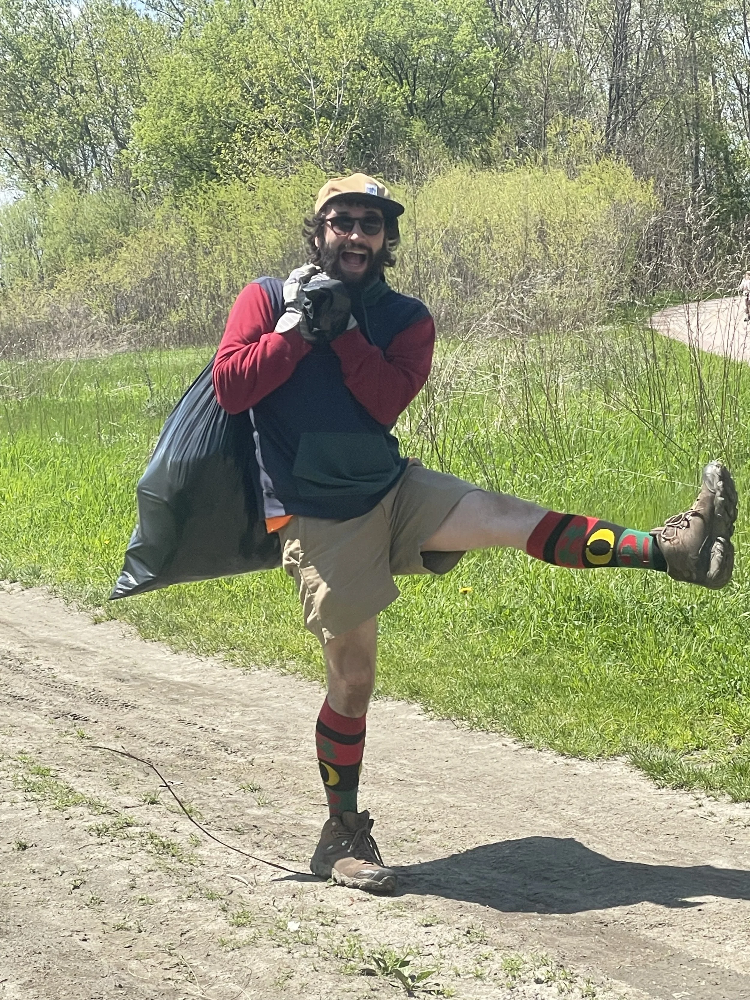 A man in outdoor attire performing a high kick on a dirt trail in a grassy area surrounded by trees, wearing sunglasses, a beige cap, and colorful socks.