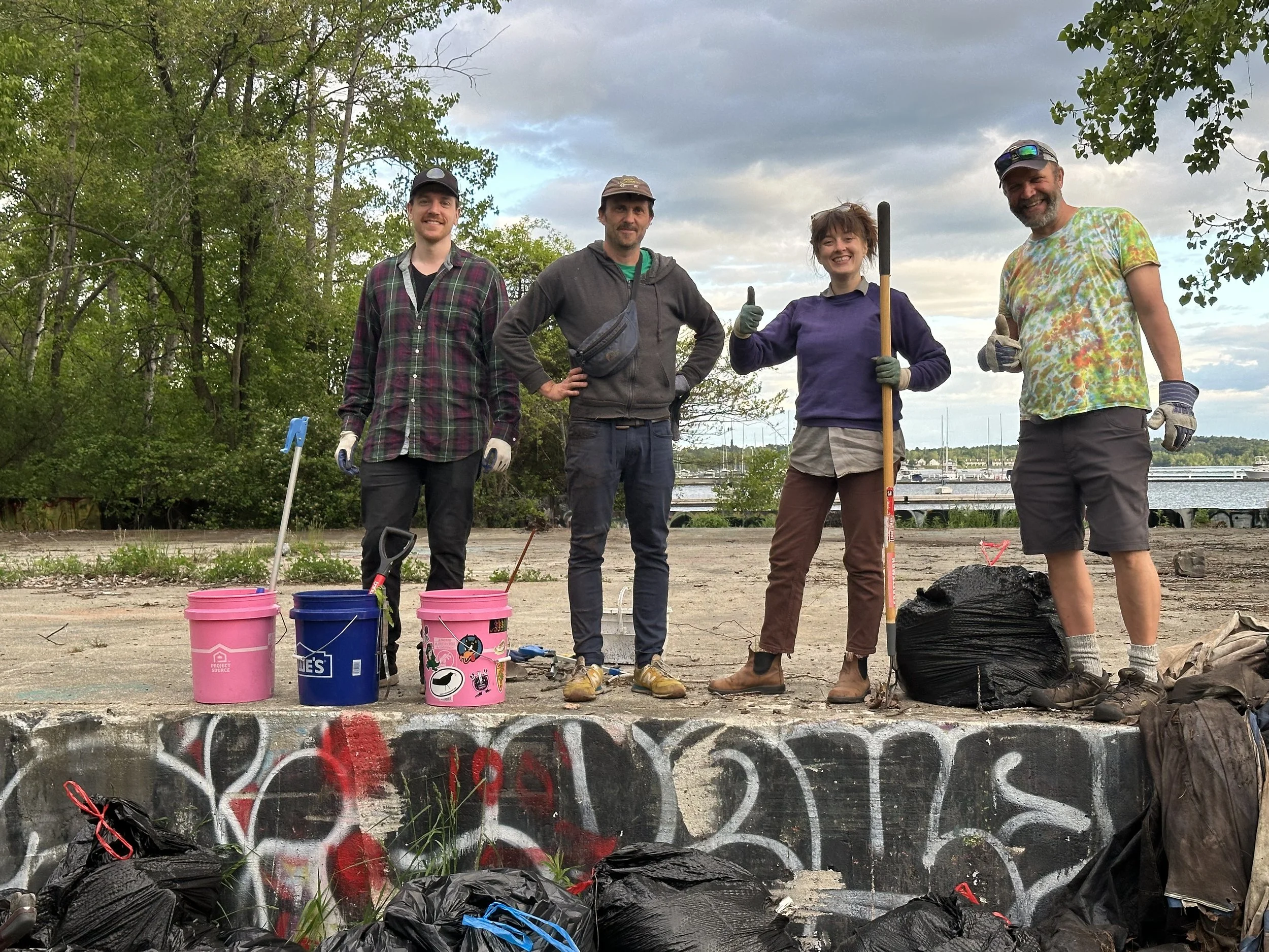 Group of f people smiling and giving thumbs up, standing outdoors along a waterfront with trees, boats, and a cloudy sky in the background. They are dressed casually, some wearing gloves, and are participating in a cleanup activity with buckets and t