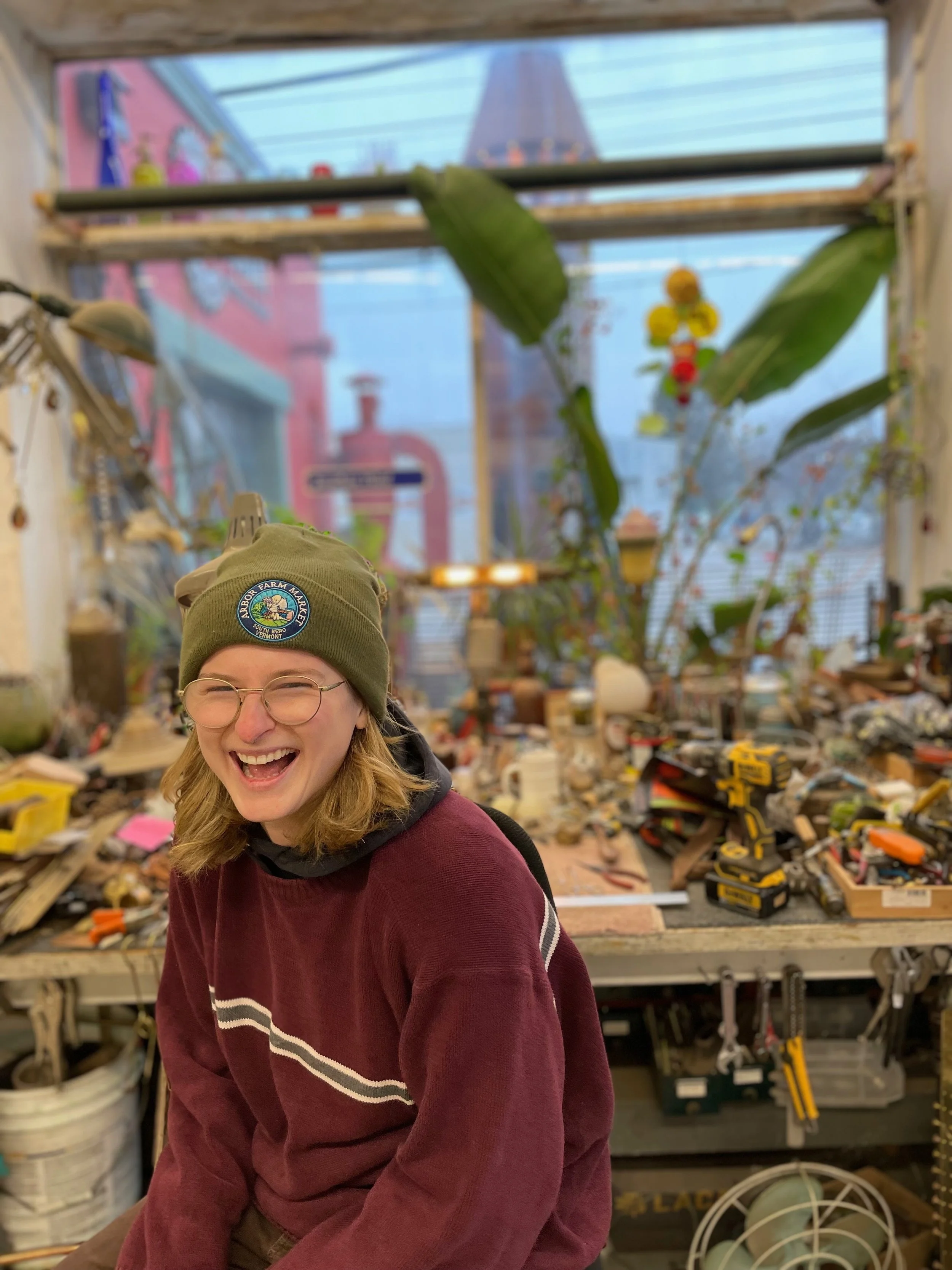 Person wearing glasses and a green beanie smiling and laughing, in a workshop or greenhouse with tools and plants in the background.