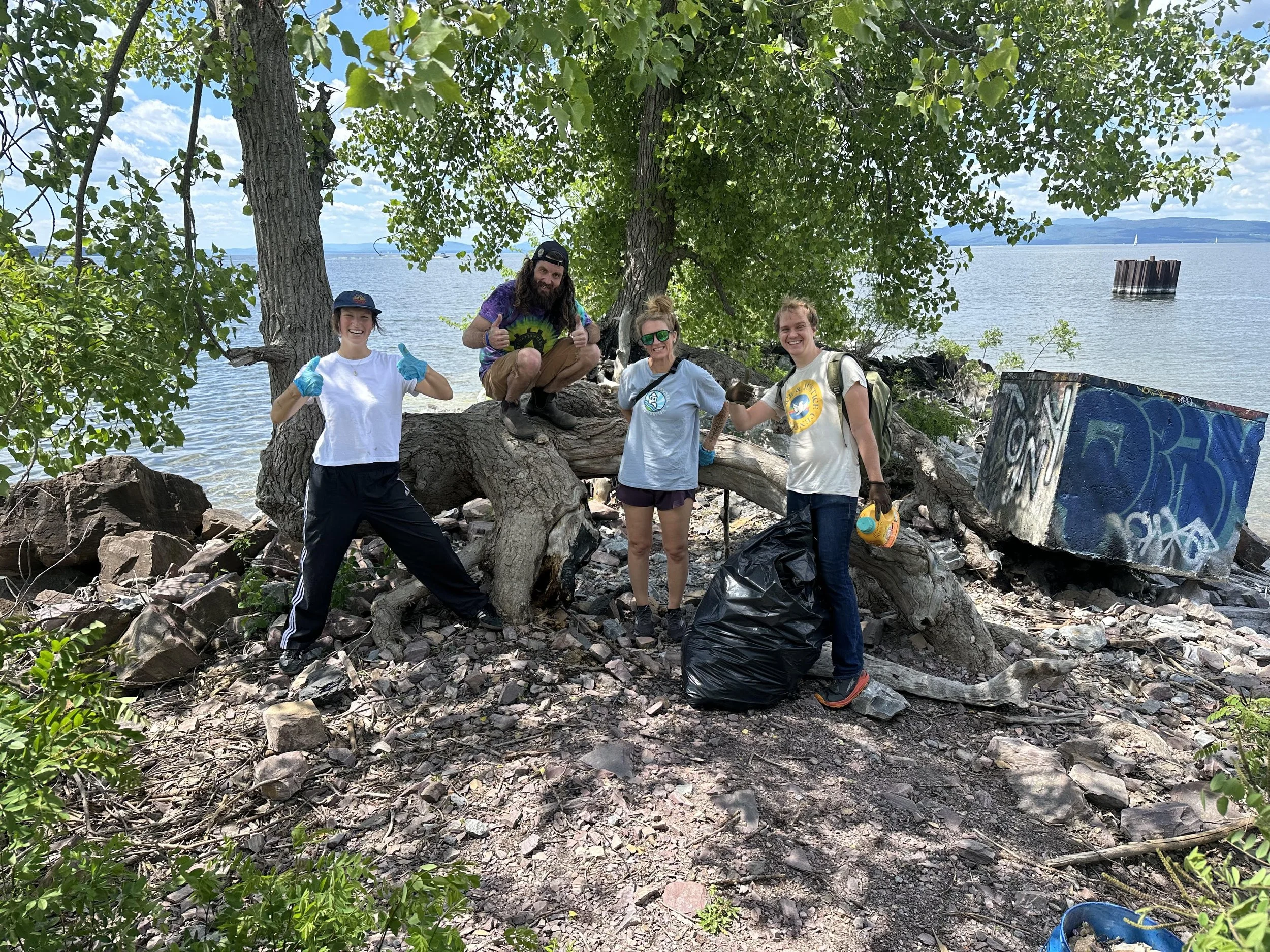 A group of five volunteers participating in a cleanup effort along a rocky shoreline by a large body of water. They are standing under a tree, smiling, and giving thumbs up. They are wearing casual clothes, gloves, and some have backpacks. There are 