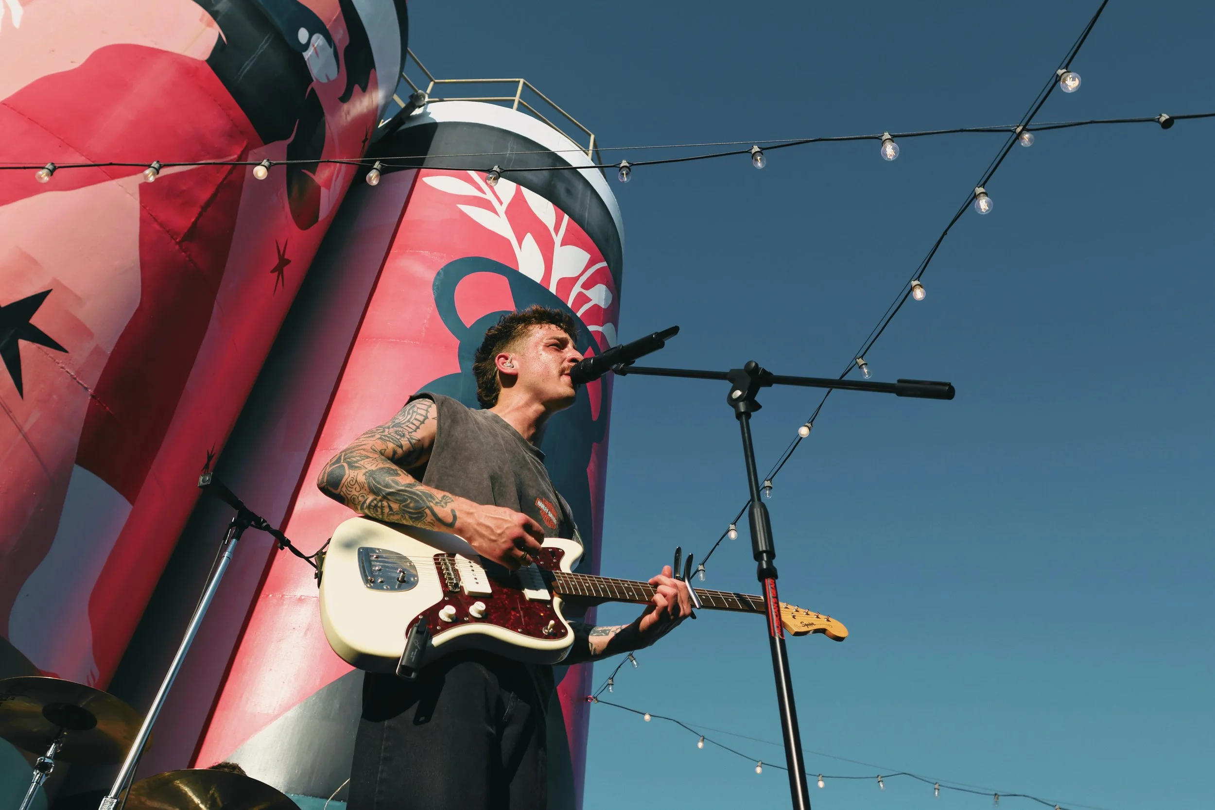 A musician with tattoos playing an electric guitar and singing into a microphone on an outdoor stage under a string of lights, with a large red and black boat or structure in the background and a clear blue sky above.