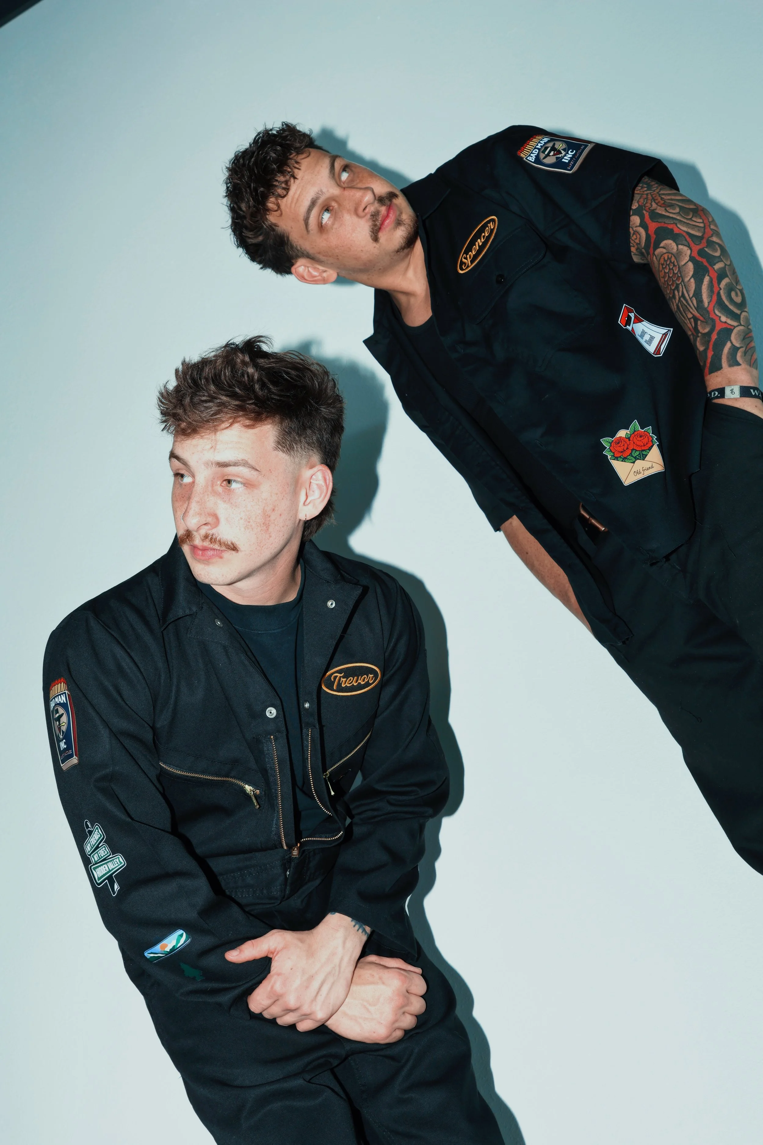 Two men in dark work jackets with patches and badges, one with a tattooed arm, standing against a plain background. One man has dark curly hair, a mustache, and is looking upward. The other man has short, dark, slightly curly hair and is looking to the side.