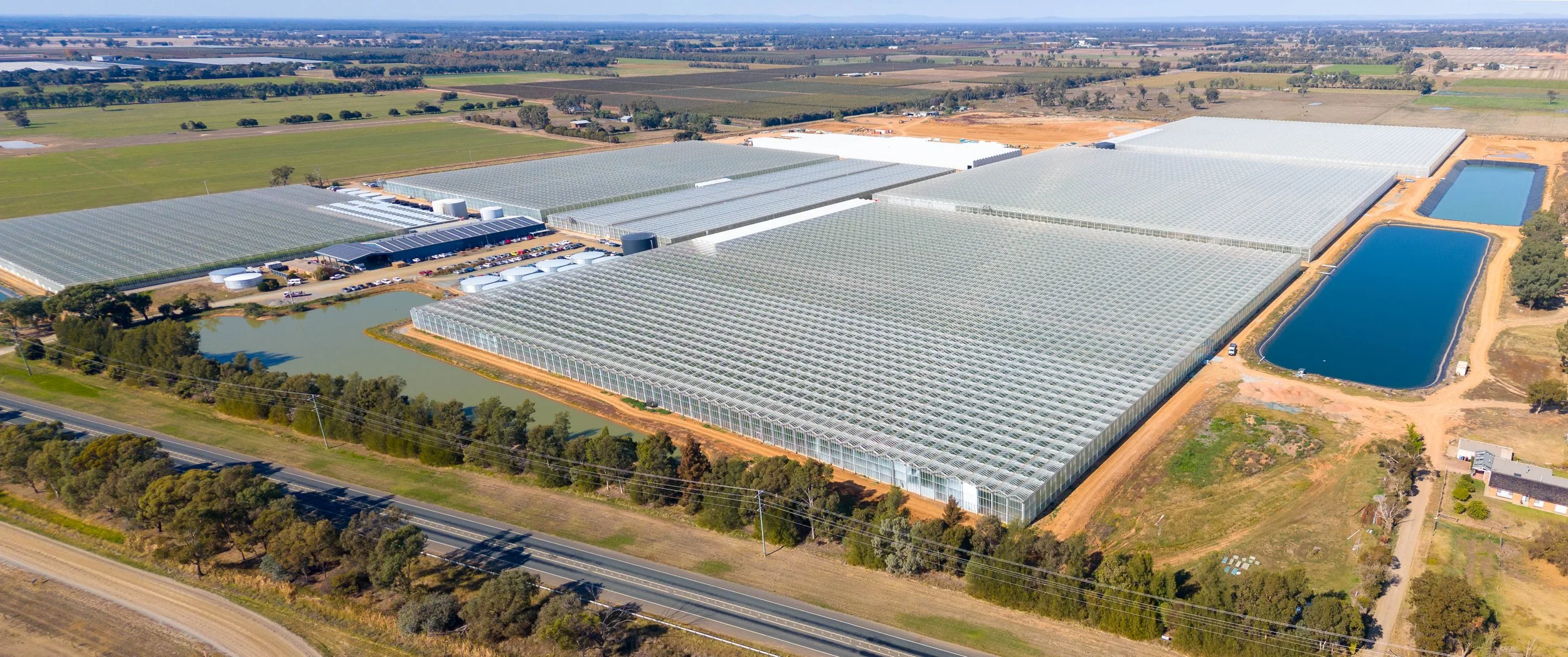 An aerial view of a large greenhouse complex with multiple greenhouses, surrounding water bodies, and nearby fields and roads.