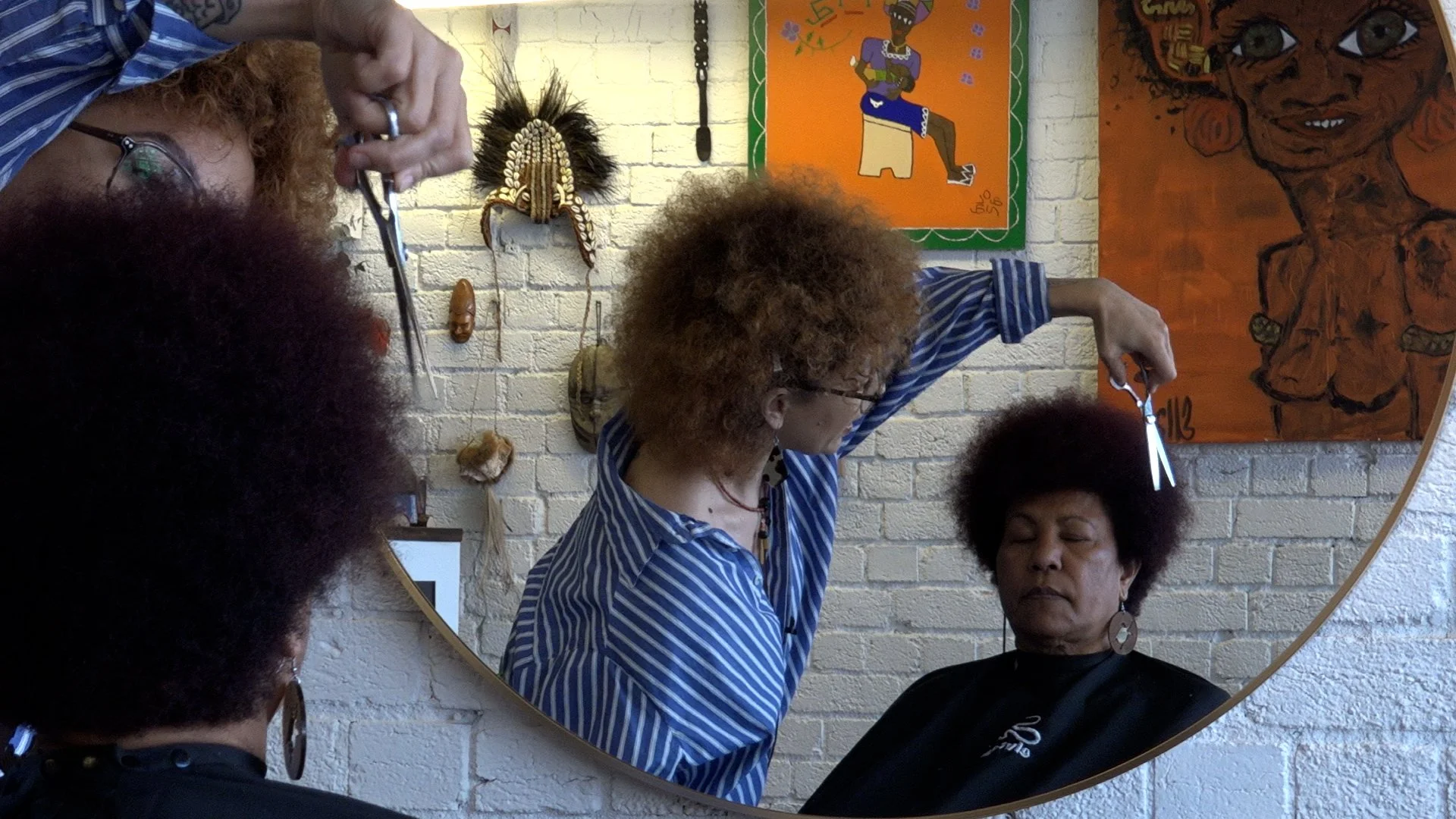 A woman with curly red hair and glasses is cutting her hair while another woman with a fro hairstyle and glasses stands nearby. In the background, there are colorful paintings and tribal masks on a white brick wall.
