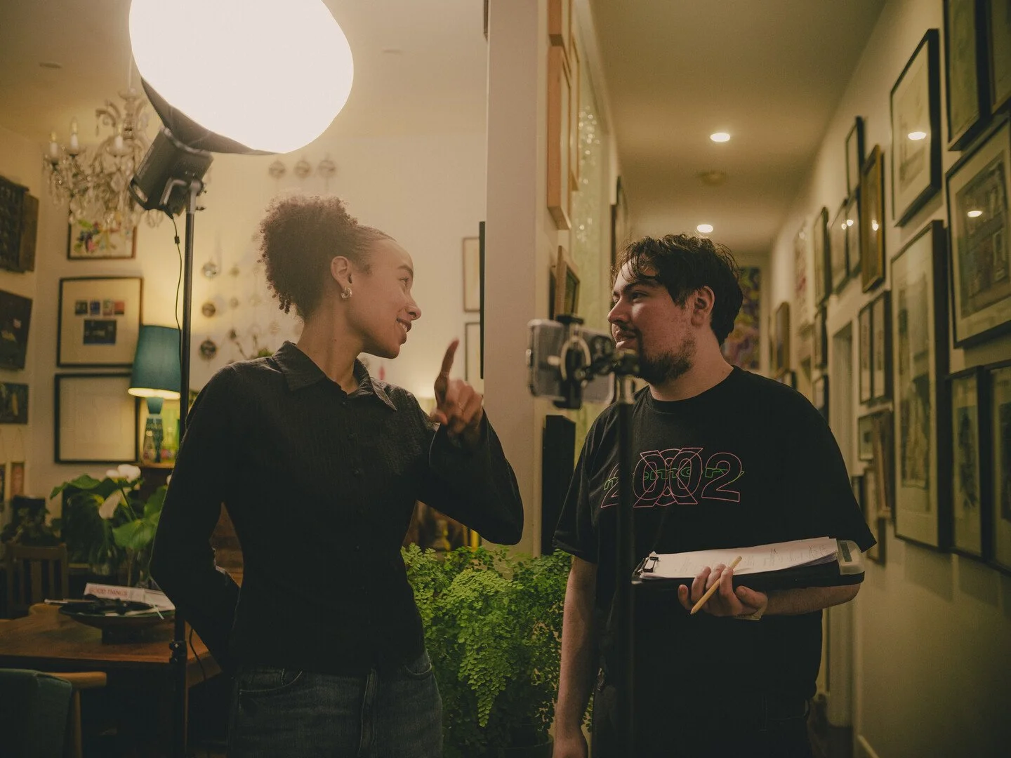 Two people having a conversation in a room with an indoor plant and framed pictures on the wall, illuminated by a large studio light.