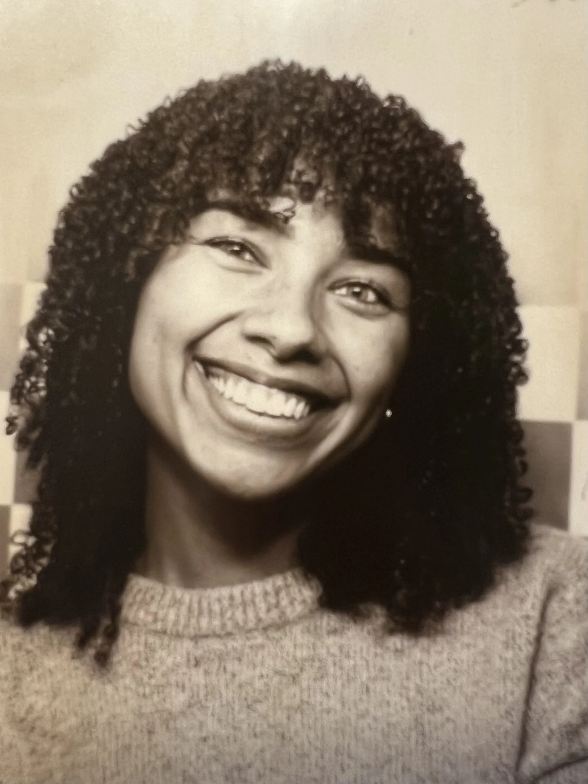 A smiling woman with curly hair wearing a light-colored knit sweater.