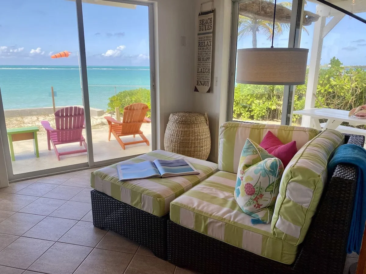 Living room with sea view, wicker basket, striped green and white couch with floral and pink pillows, sliding glass door leading to outdoor patio with colorful Adirondack chairs and ocean scenery.