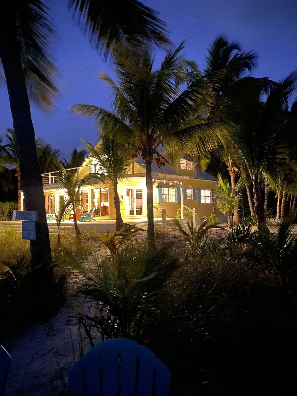 A beach house illuminated at night surrounded by palm trees, with lights shining through the windows and outdoor seating visible on the porch.