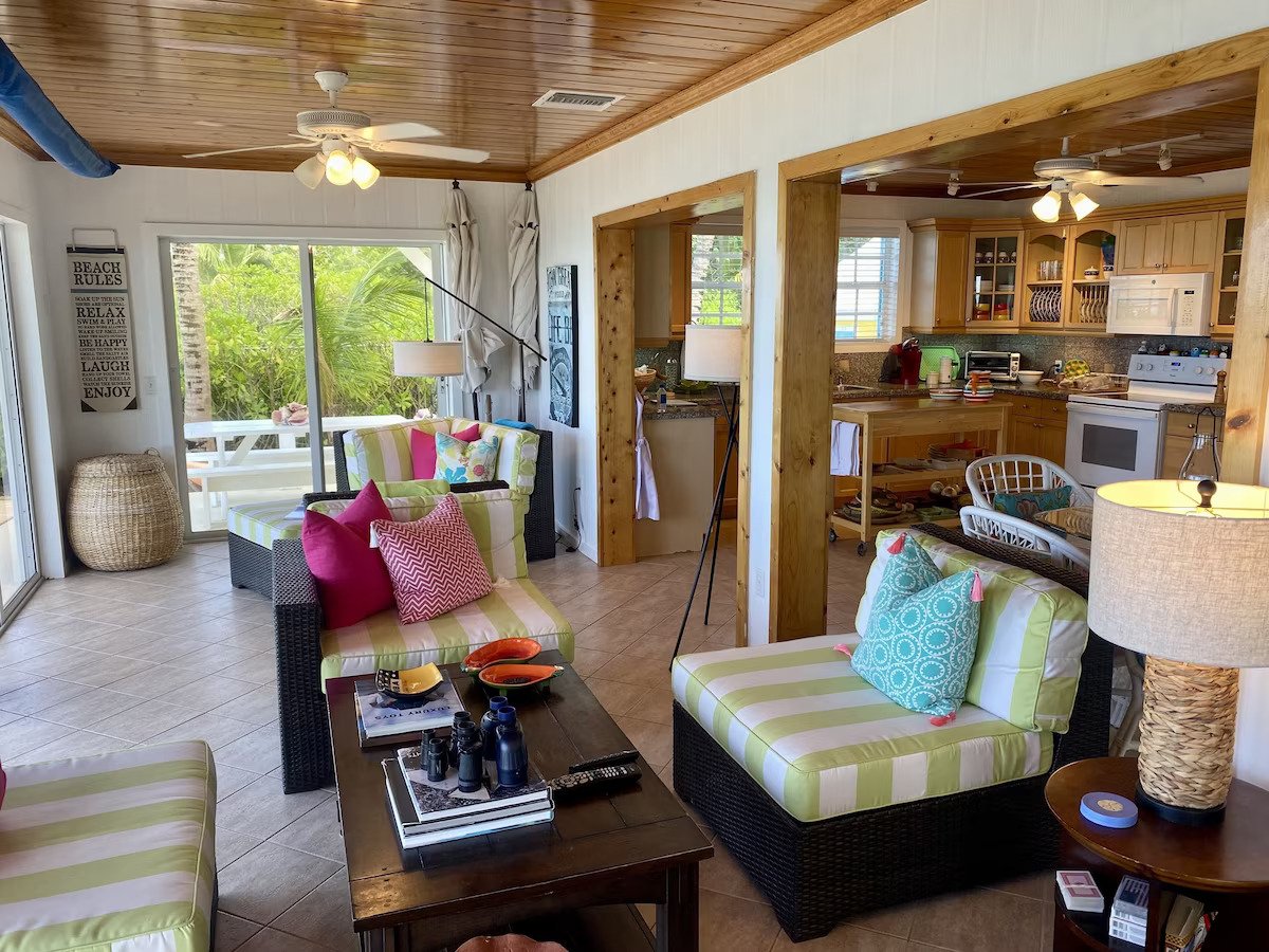 Living room with wicker furniture featuring green and white striped cushions, pink and teal pillows, a wooden coffee table, and a view of the kitchen with wooden cabinets and white appliances.