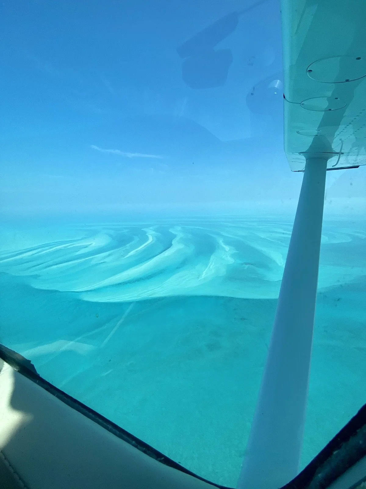 View from inside an aircraft showing clear blue water and sky, with the aircraft's wing visible through the window.