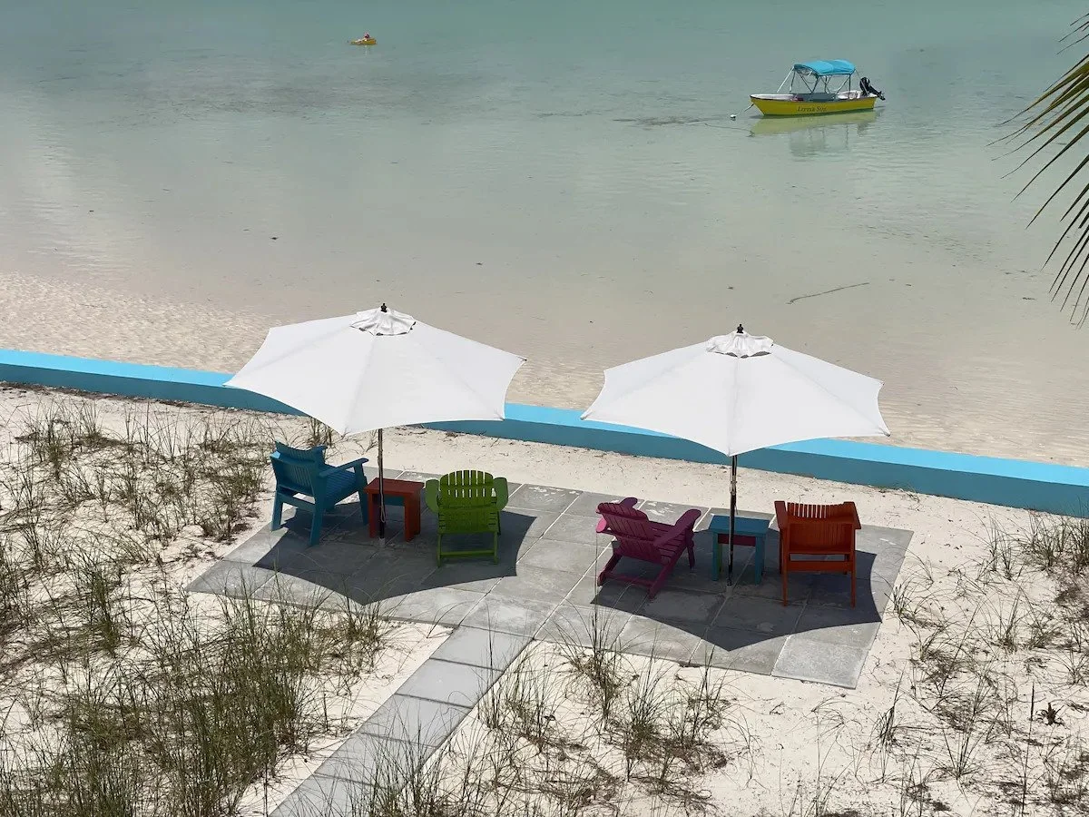 Two white beach umbrellas over colorful chairs and tables on a paved area by the beach, with boats floating on calm water in the background.