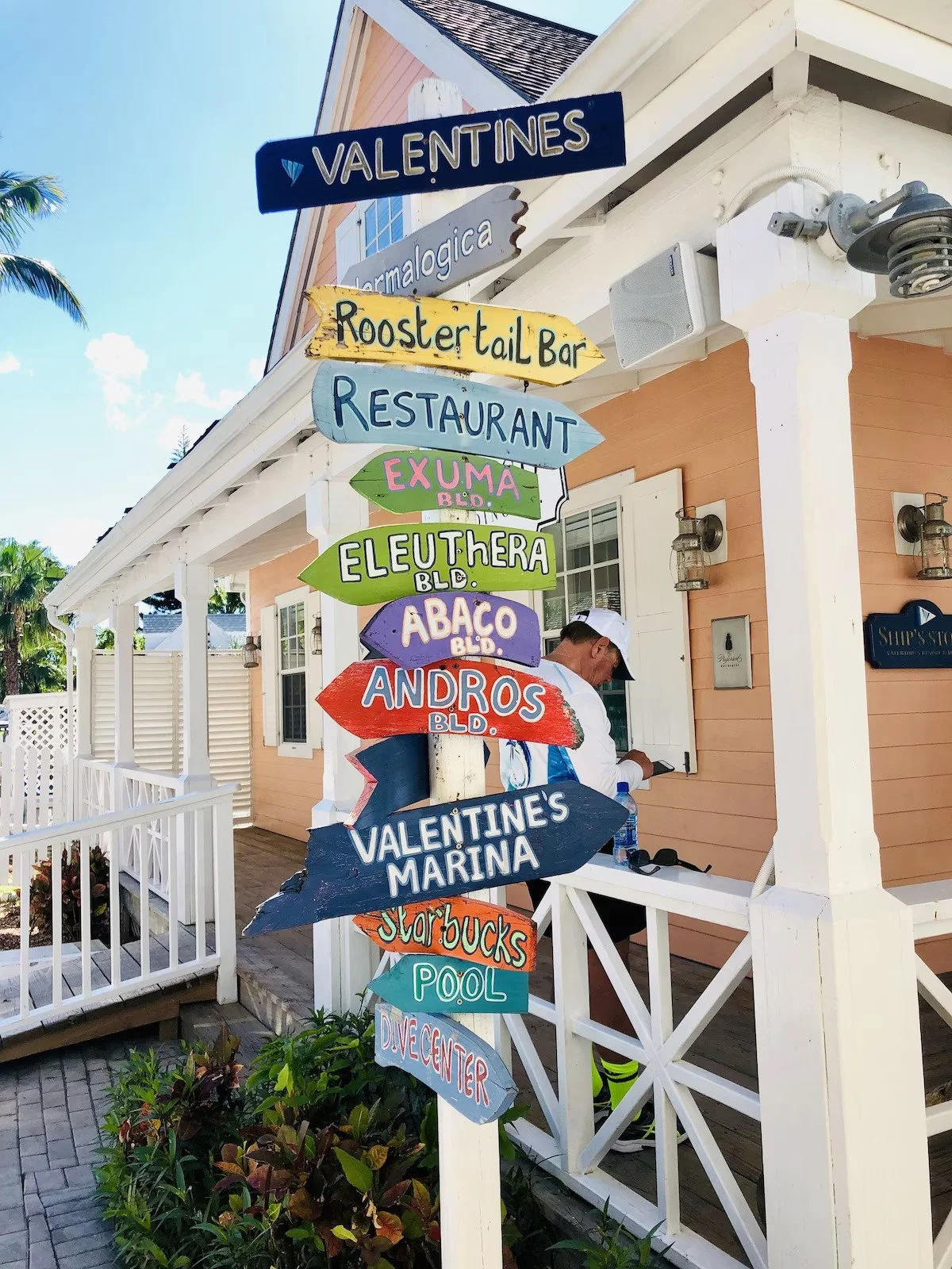 Colorful directional signs on a white post outside a building with a man in a white cap and blue shirt checking his phone. The signs point to various locations such as Valentines, Rooster Tail Bar, Restaurant, Exuma, Eleuthera, Abaco, Andros, Valentines Marina, and Pool, with a blue sky and palm trees in the background.