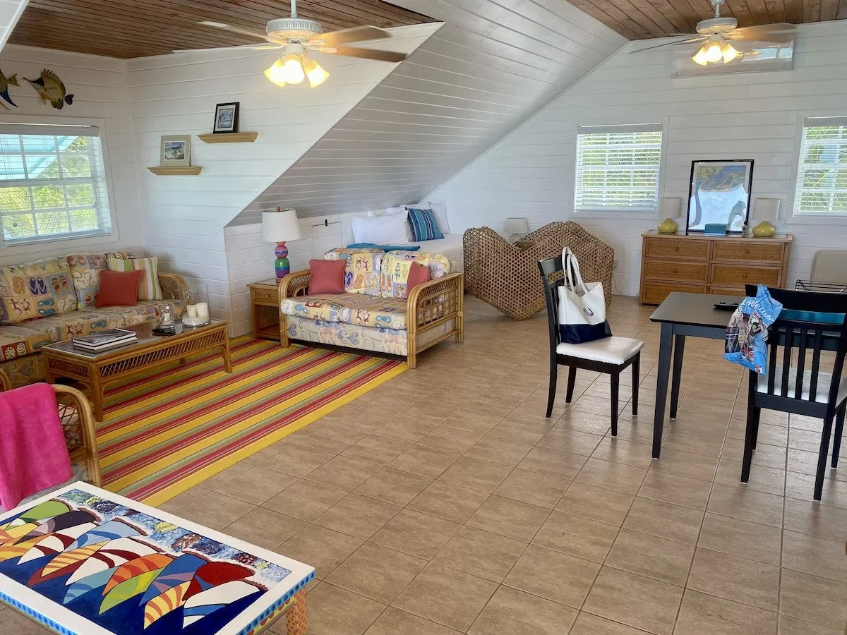 Spacious living room with white panel walls, tiled floor, rustic ceiling, ceiling fans, and beach-themed decor. Contains wicker and upholstered furniture, a colorful striped rug, a black dining table with chairs, and various decorative items on furni