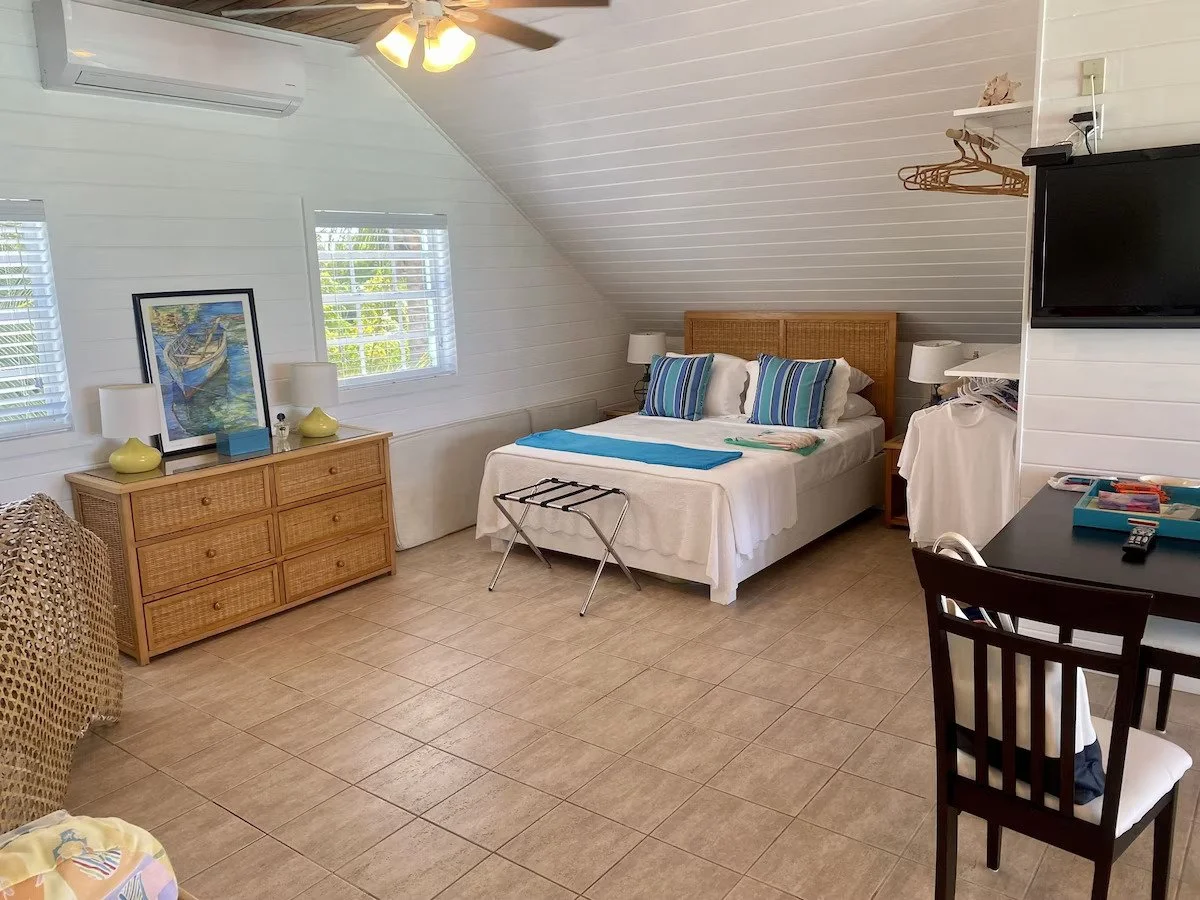 A cozy bedroom with white paneled walls and ceiling, featuring a queen bed with striped pillows, a wooden headboard, and two nightstands with lamps. There is a wicker dresser with a framed picture and lamps, a wall-mounted TV, and a small table with 