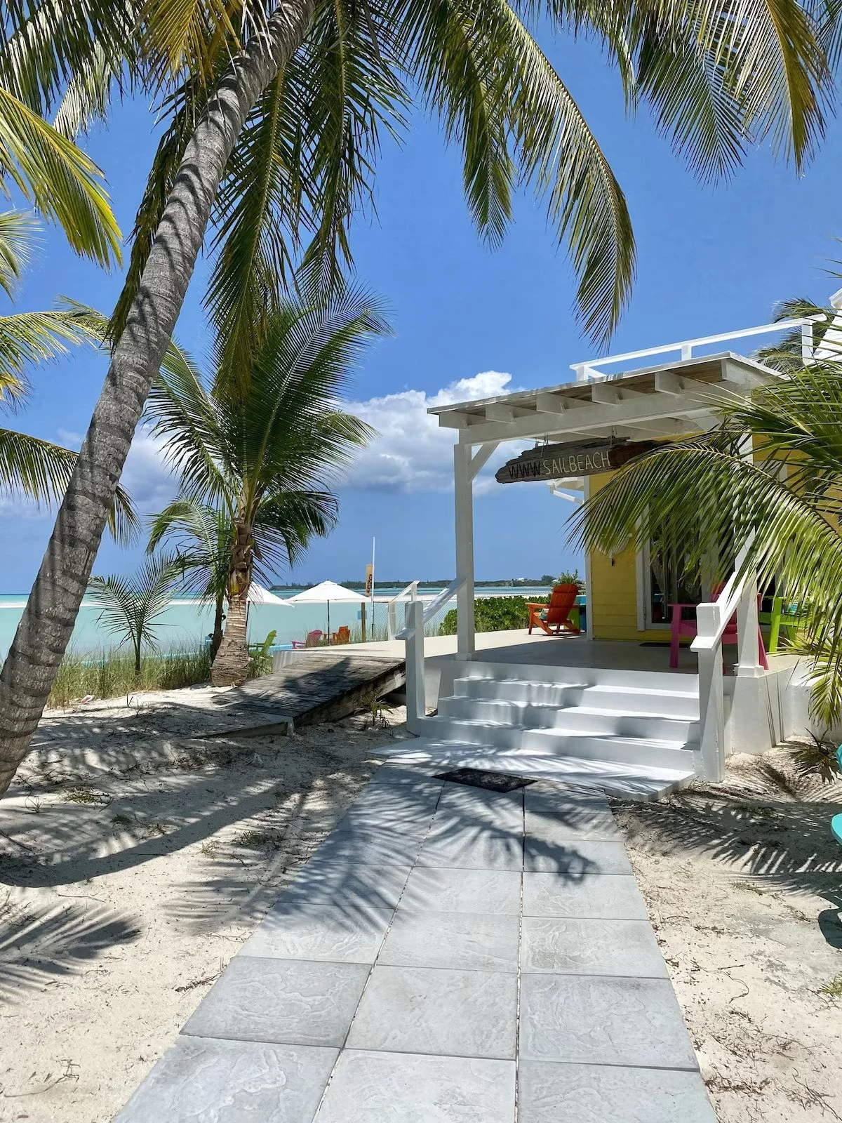 Beachside scene with palm trees, a white cottage with stairs, colorful chairs, umbrellas, and ocean in the background.