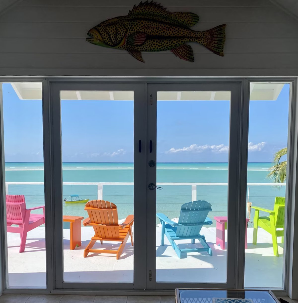 View of the ocean through sliding glass doors with colorful outdoor chairs and tables on a balcony, and a fish decoration above the door.