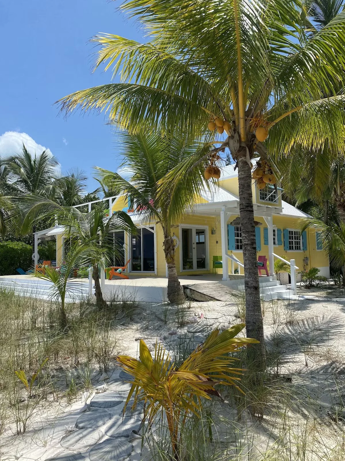 A yellow beach house with white trim, blue shutters, and a front porch, surrounded by palm trees on a sandy beach under a clear blue sky.