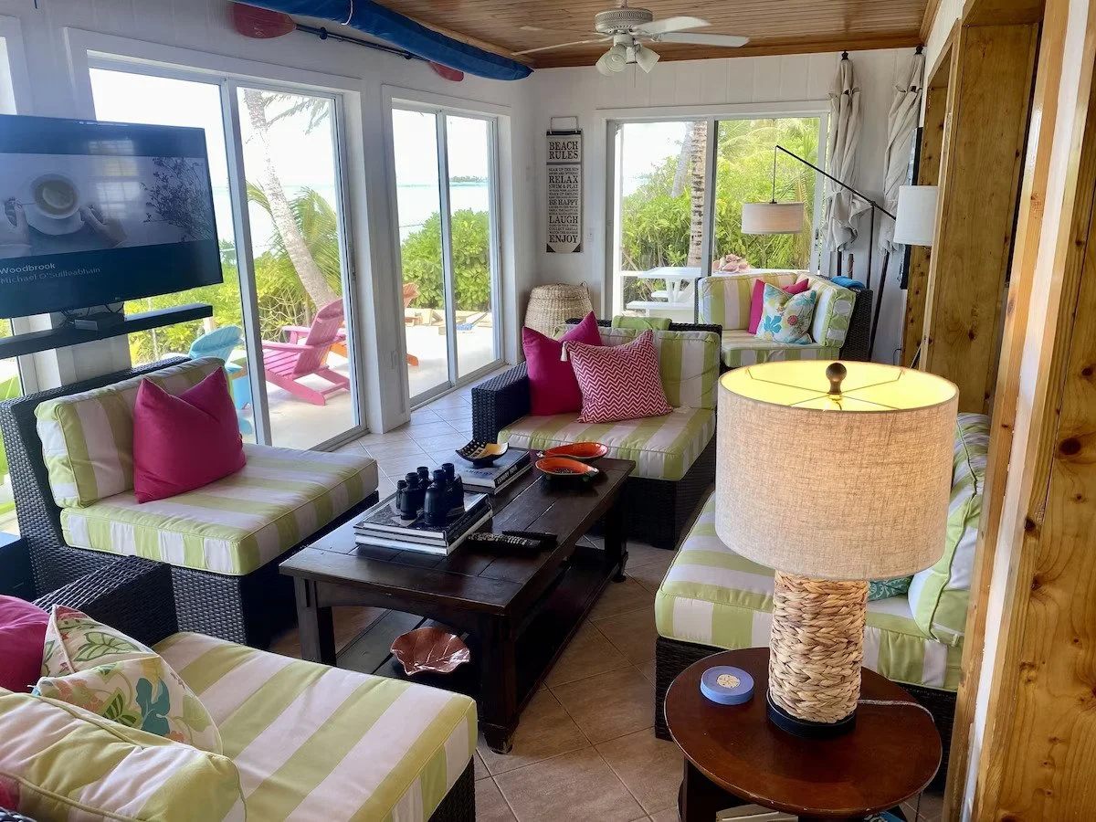 Living room with tropical decor, wicker furniture with striped green and white cushions, colorful pillows, a wooden coffee table with binoculars and remote controls, a lamp with a woven base, and sliding glass doors leading to an outdoor patio with p