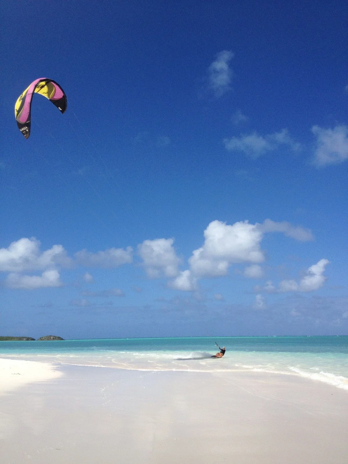 A person kiteboarding on a beach with a blue sky and scattered clouds, turquoise water in the background, and white sand in the foreground.