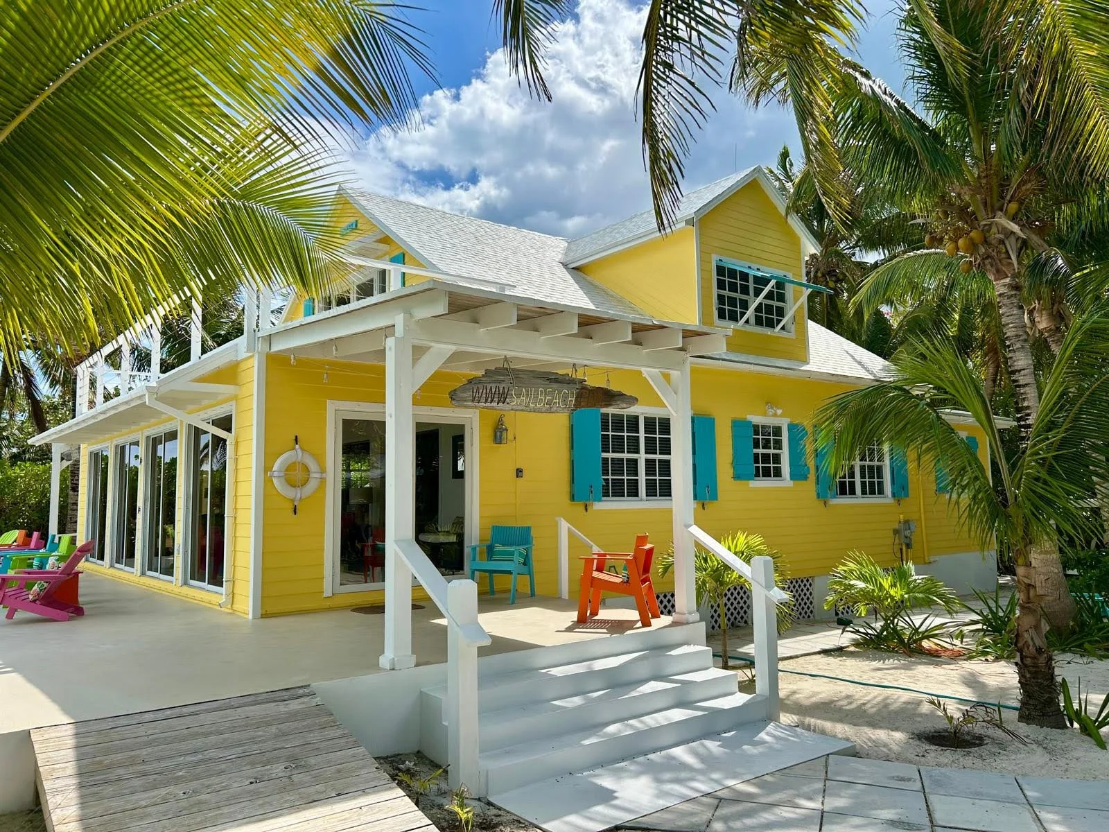 Colorful yellow beach house with turquoise shutters, surrounded by palm trees, with a porch, outdoor chairs, and a sign reading 'www.sanbeach,' under a partly cloudy sky.