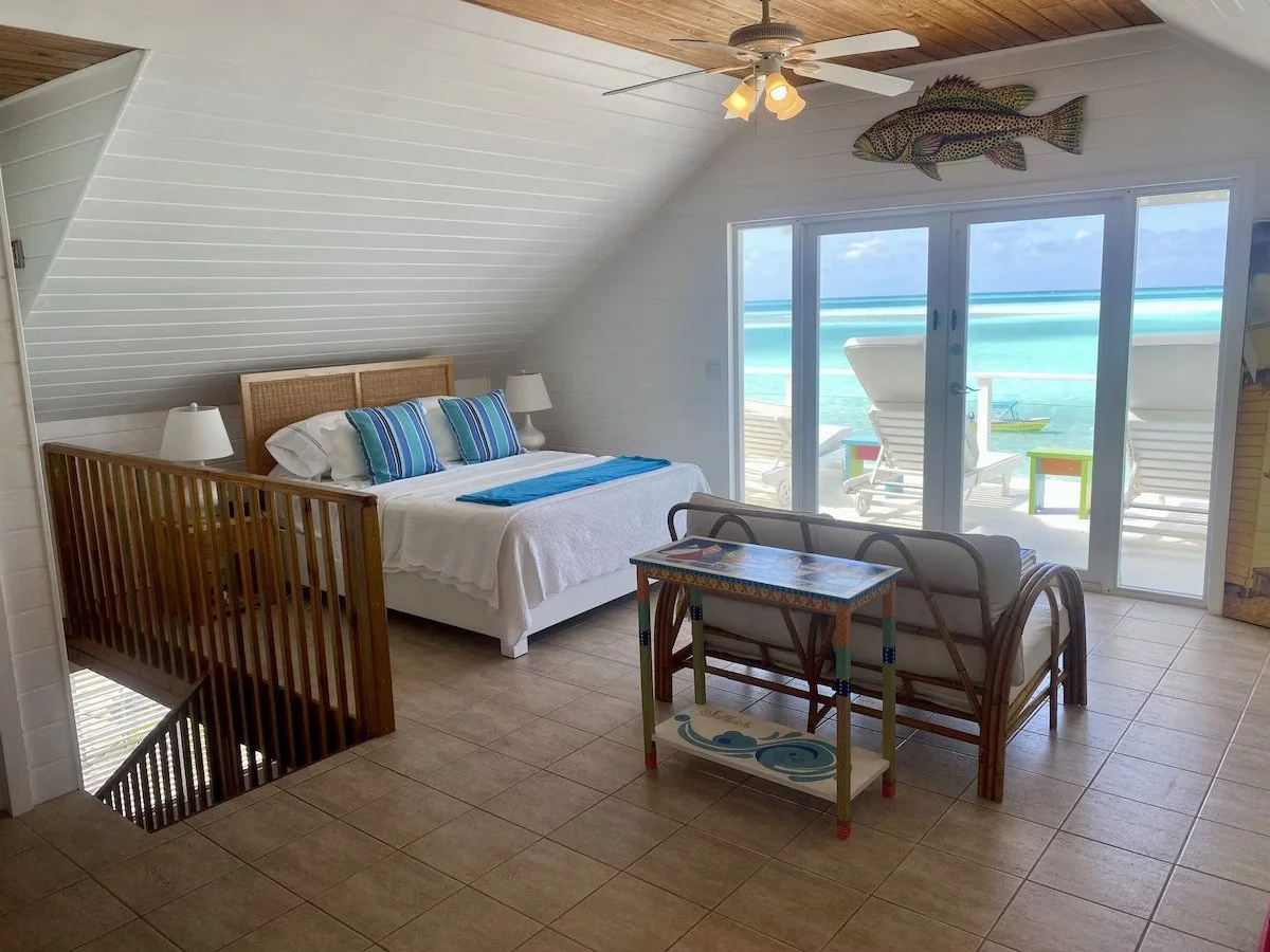 Beachside bedroom with white walls and ceiling, a bed with striped pillows, sliding glass door leading to a balcony with ocean view, and decorative fish wall art.