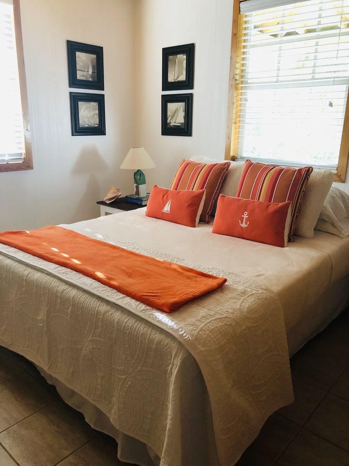 A cozy bedroom with a white quilted bedspread, orange towels and pillows with nautical symbols, striped pillows, a nightstand with a lamp, a seashell, and a book, and framed sailboat pictures on the wall. The room has light-colored walls and windows with blinds.
